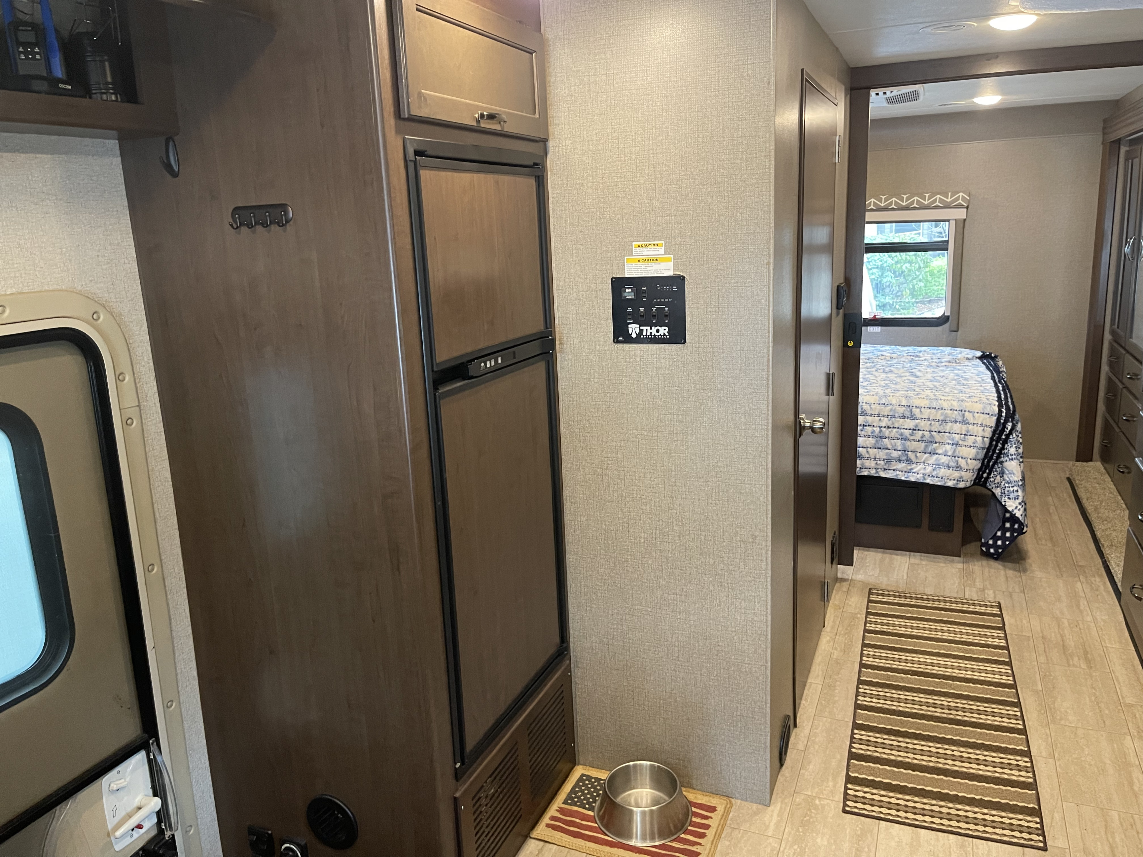 Inside an RV rental: a modern kitchen area with fridge & control panel, leading to a cozy bedroom. Includes a pet bowl.. Thor Freedom Traveler 2020