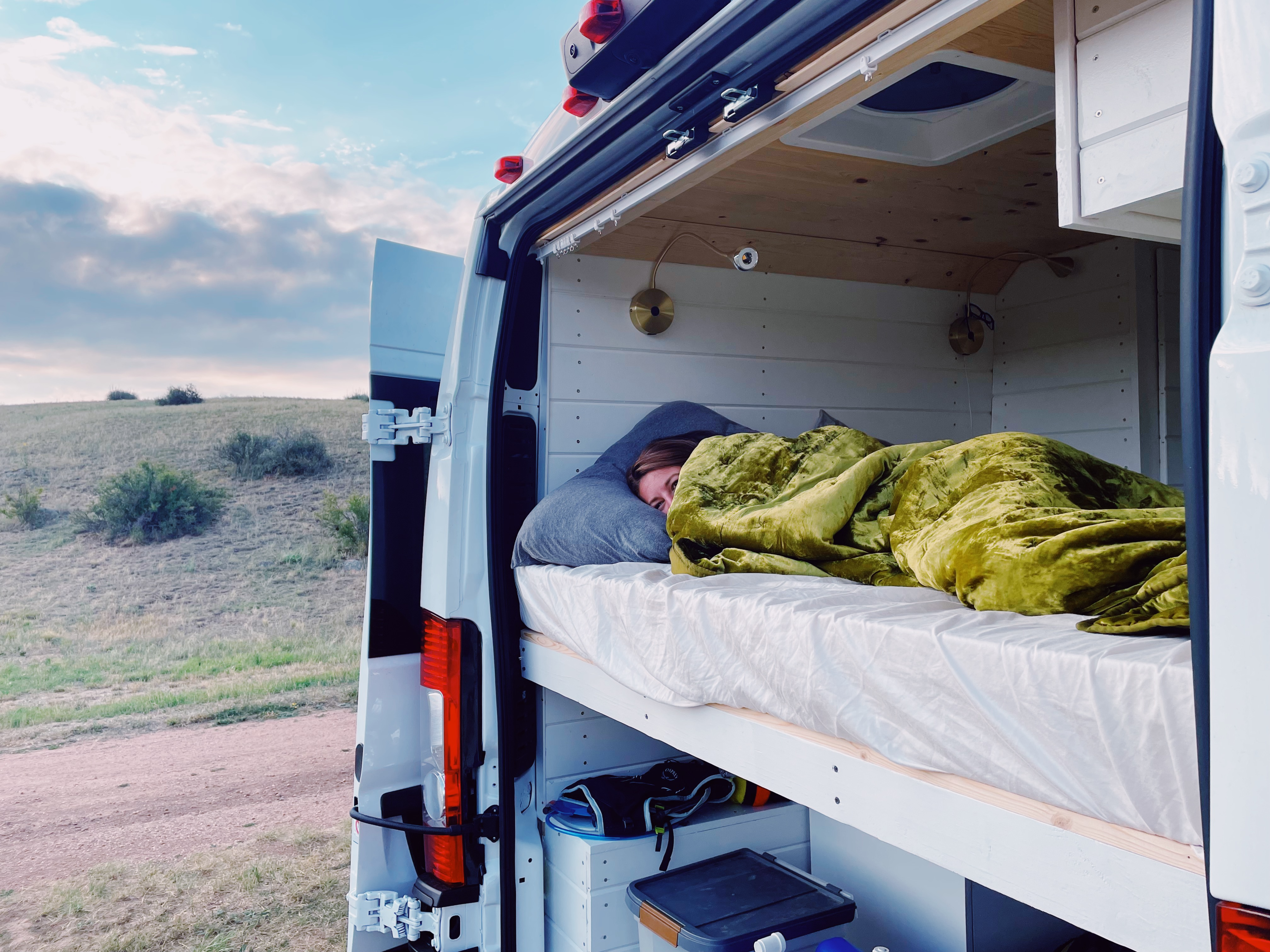 A person is sleeping under a green blanket in a white RV rental van, with the back doors open, revealing a grassy hillside.. Dodge Promaster 2500 2021