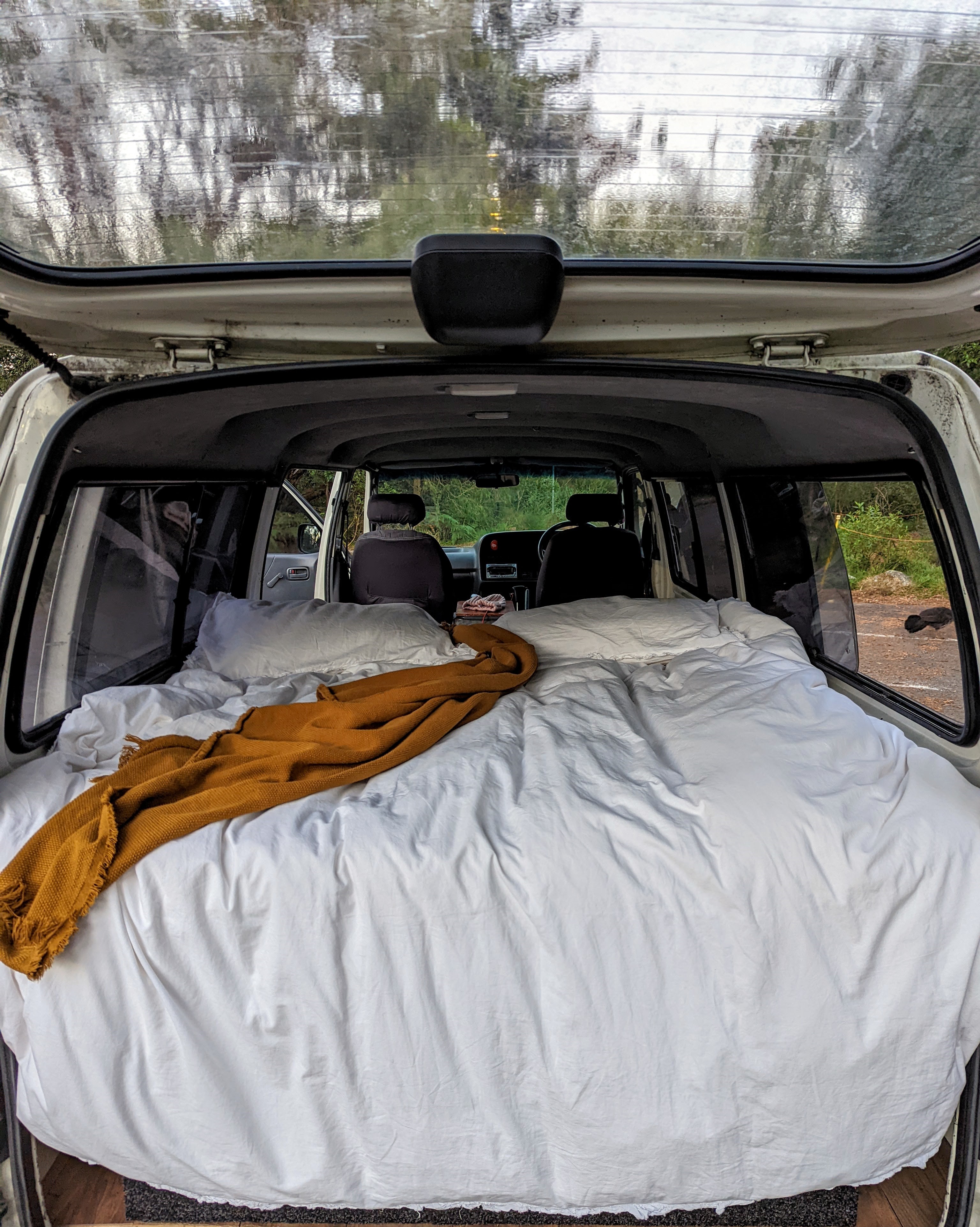 Cozy campervan bed setup in a parked RV rental, with a view through the front window to green trees outside.. Toyota HiAce 2003