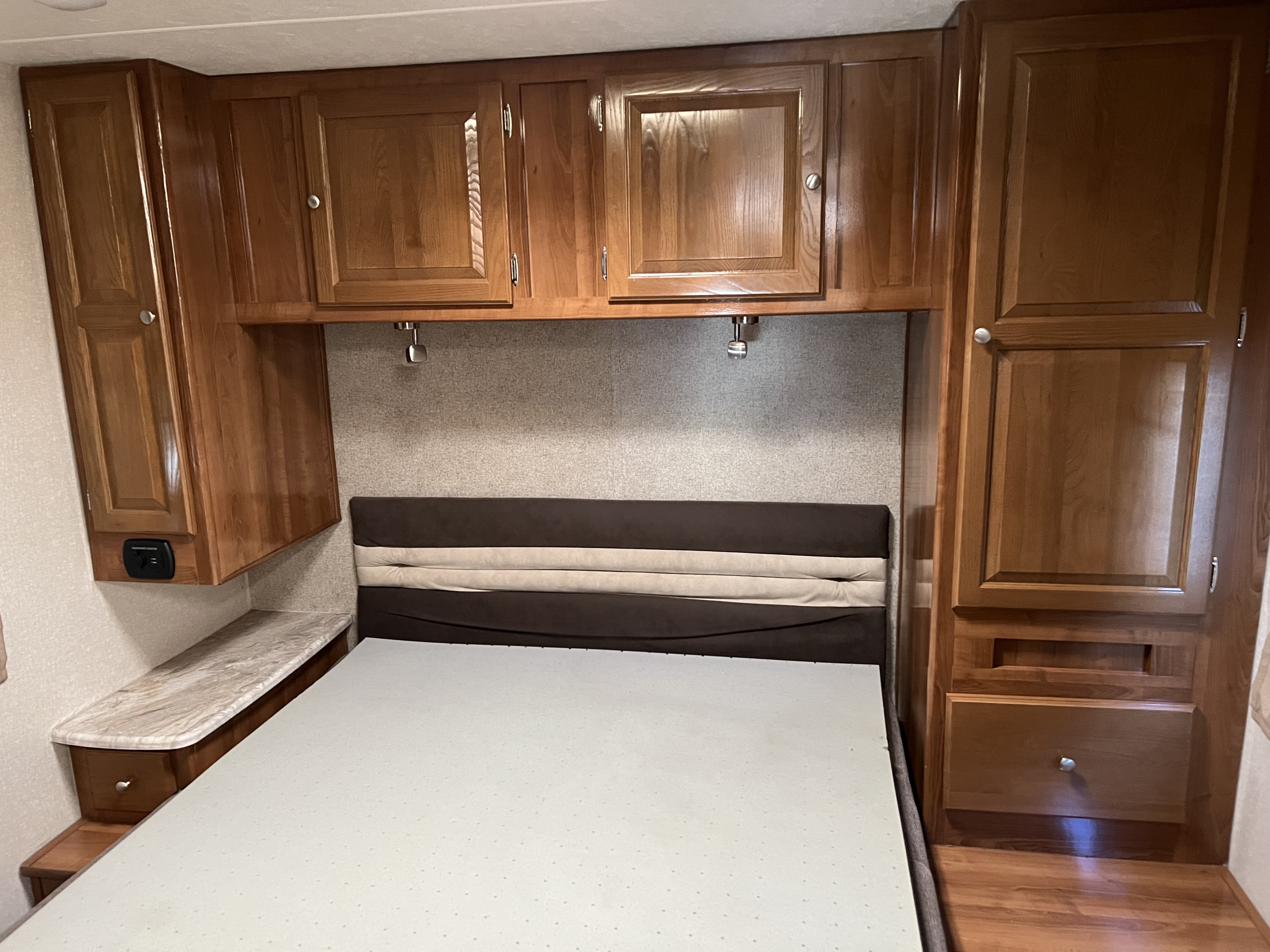 RV rental bedroom with wooden overhead cabinets, an upholstered bed, bedside table, and charging port.. Coachmen Leprechaun 2019