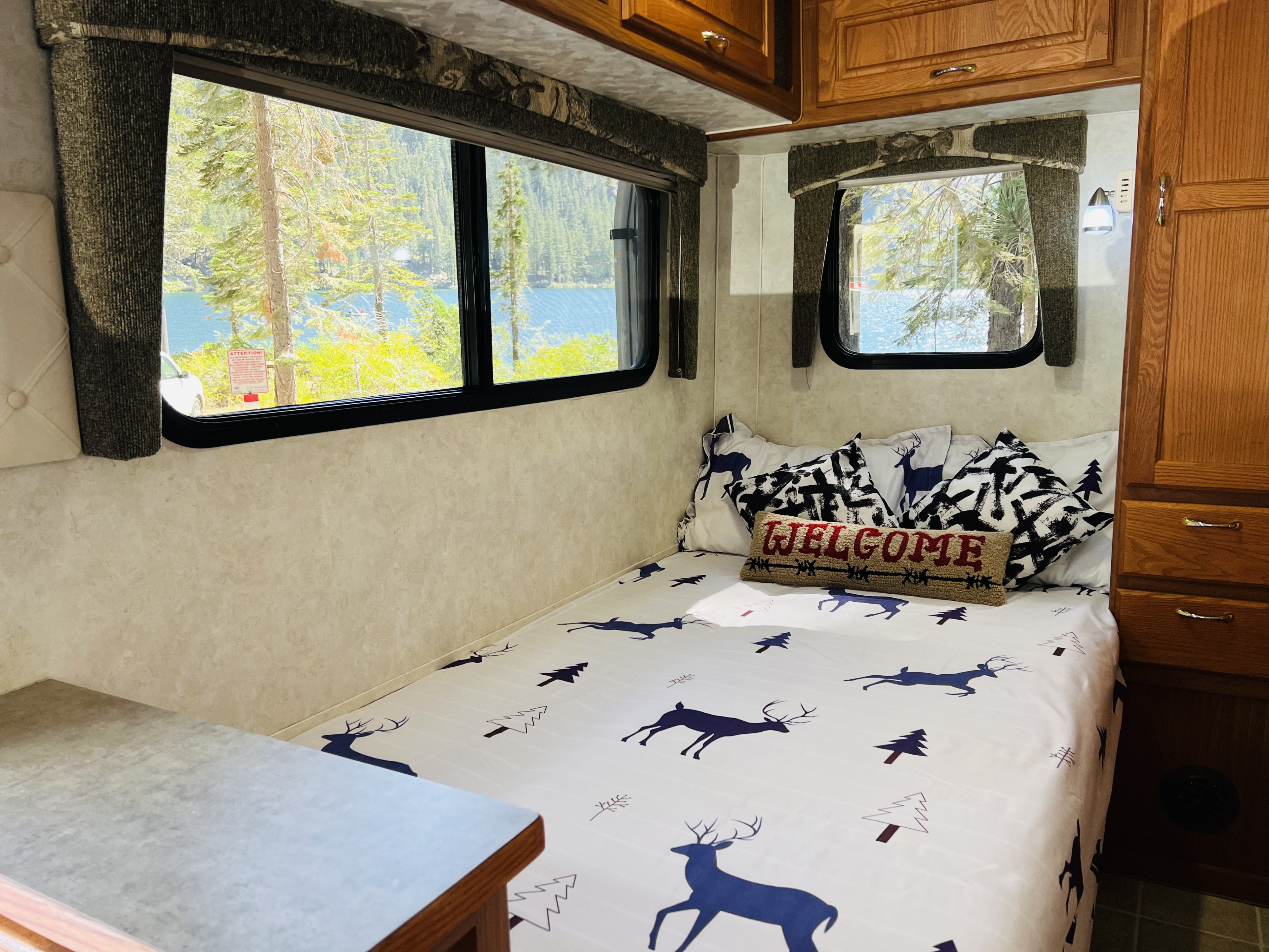 Cozy RV rental bedroom with a large window showcasing a stunning lake and mountain view, complete with deer-patterned bedding.. Bigfoot RV Bigfoot RV Trailer 2005
