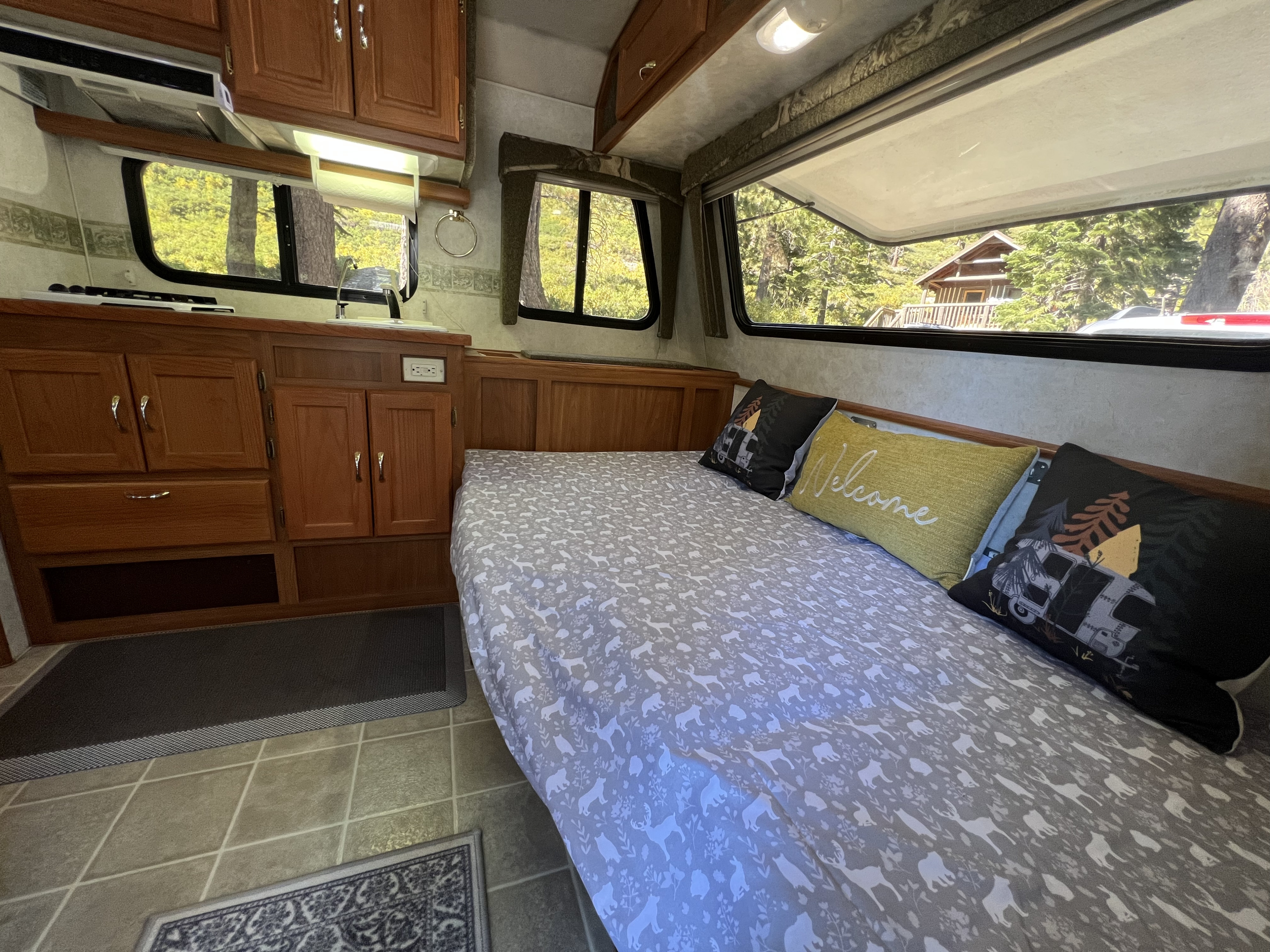 Inside a cozy RV rental: kitchen, comfortable bed with forest-themed bedding, and expansive windows showing nature views.. Bigfoot RV Bigfoot RV Trailer 2005