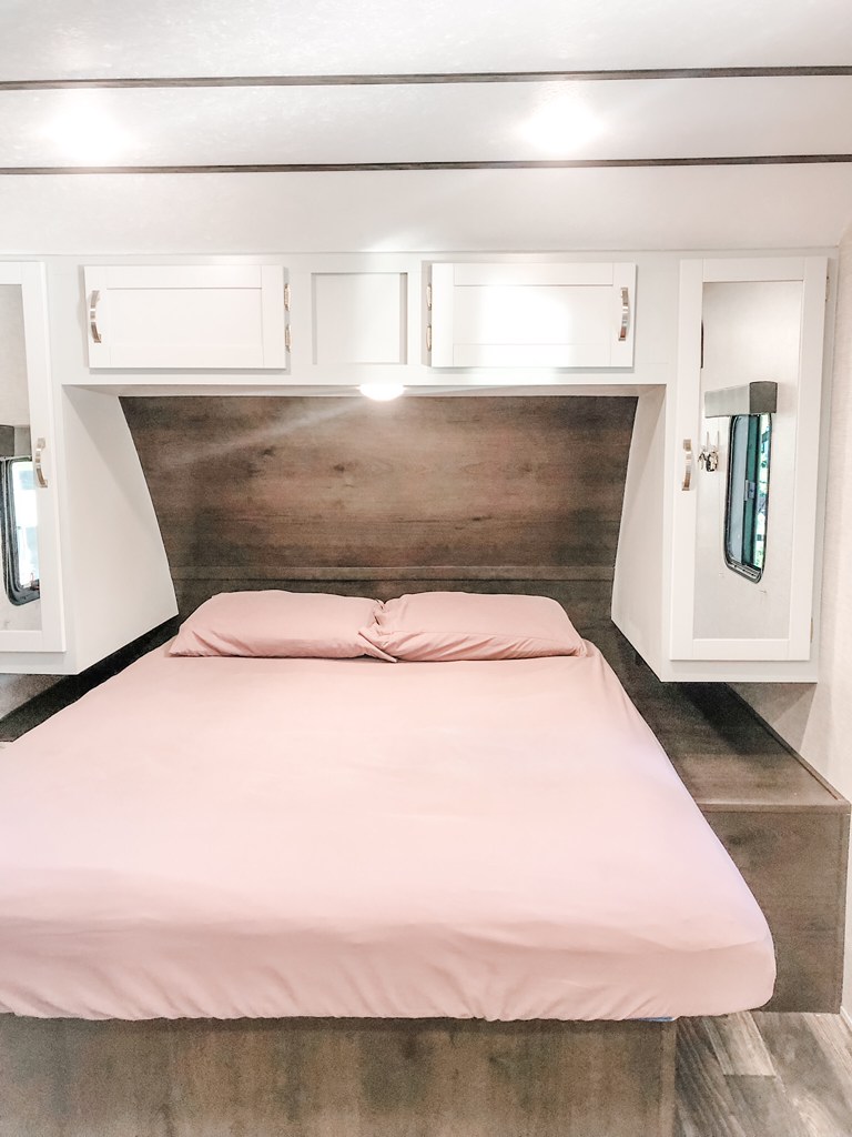 Clean, modern RV rental bedroom with soft pink bedding, white overhead cabinets, and a wood headboard. Cozy travel space.. Keystone RV Bullet Ultra Lite 2019
