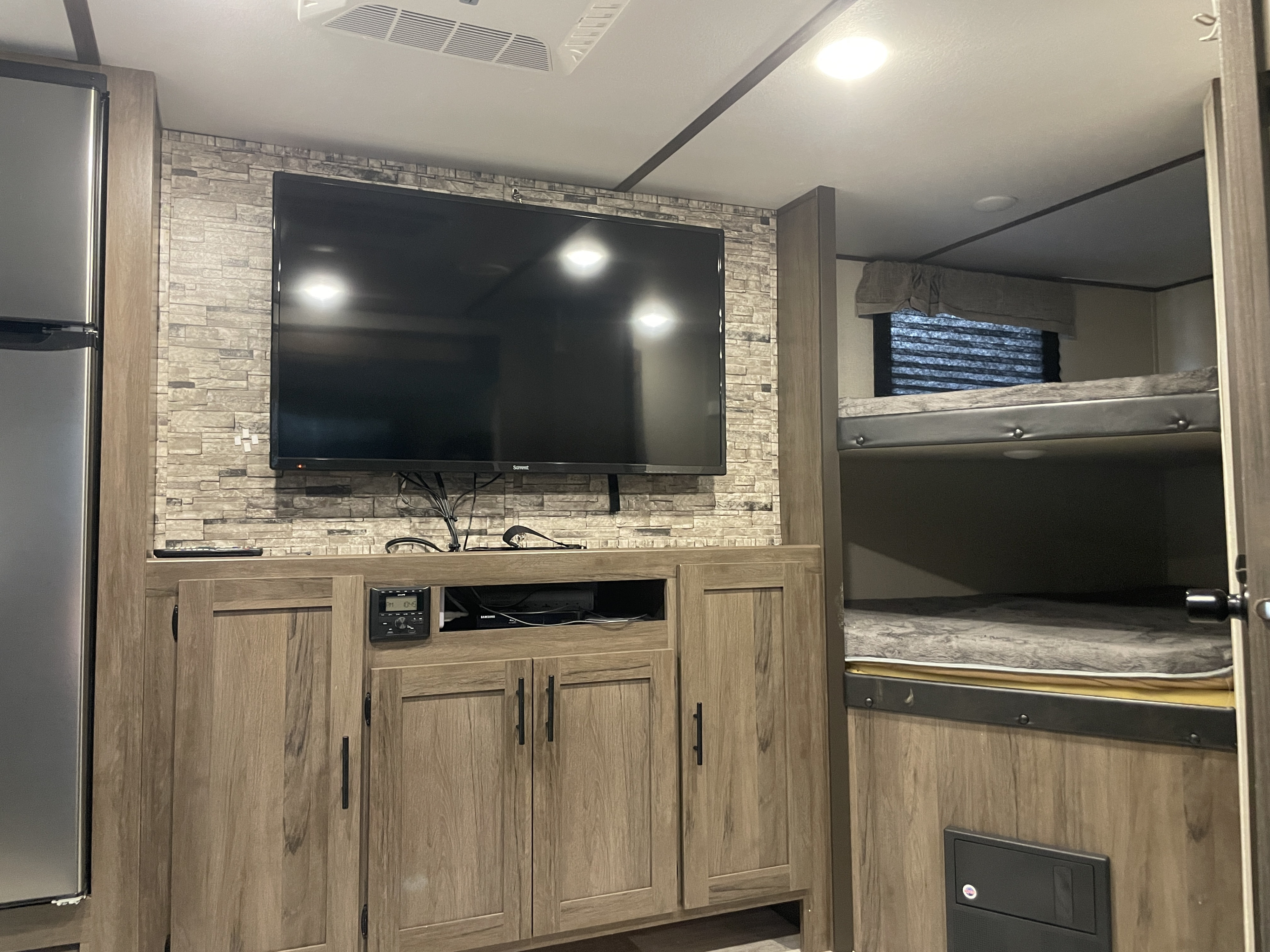 Spacious RV rental interior with a TV entertainment center on a stone accent wall, refrigerator, and built-in bunk beds.. Prime Time Avenger 2020