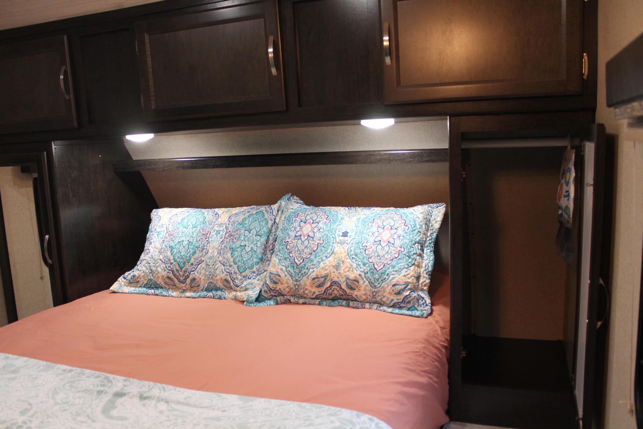 Comfortable RV rental bedroom with a bed, colorful patterned pillows, peach sheets, dark wood cabinets, lights & storage.. Coachmen Spirit Ultralight 2020