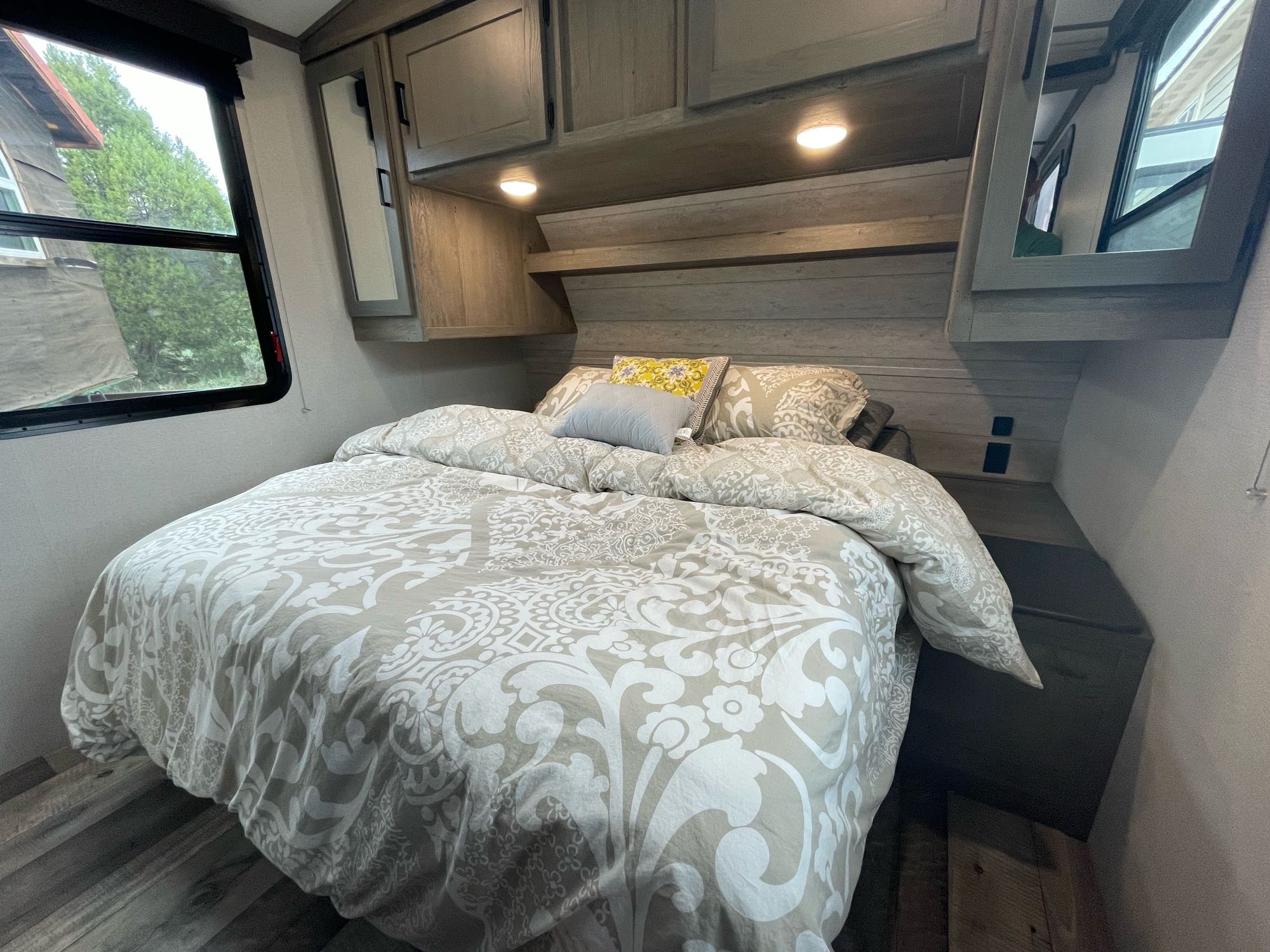 Comfortable RV rental bedroom featuring a large bed with a patterned duvet, overhead cabinets, and a window overlooking greenery.. Keystone RV Outback Ultra-Lite 2021