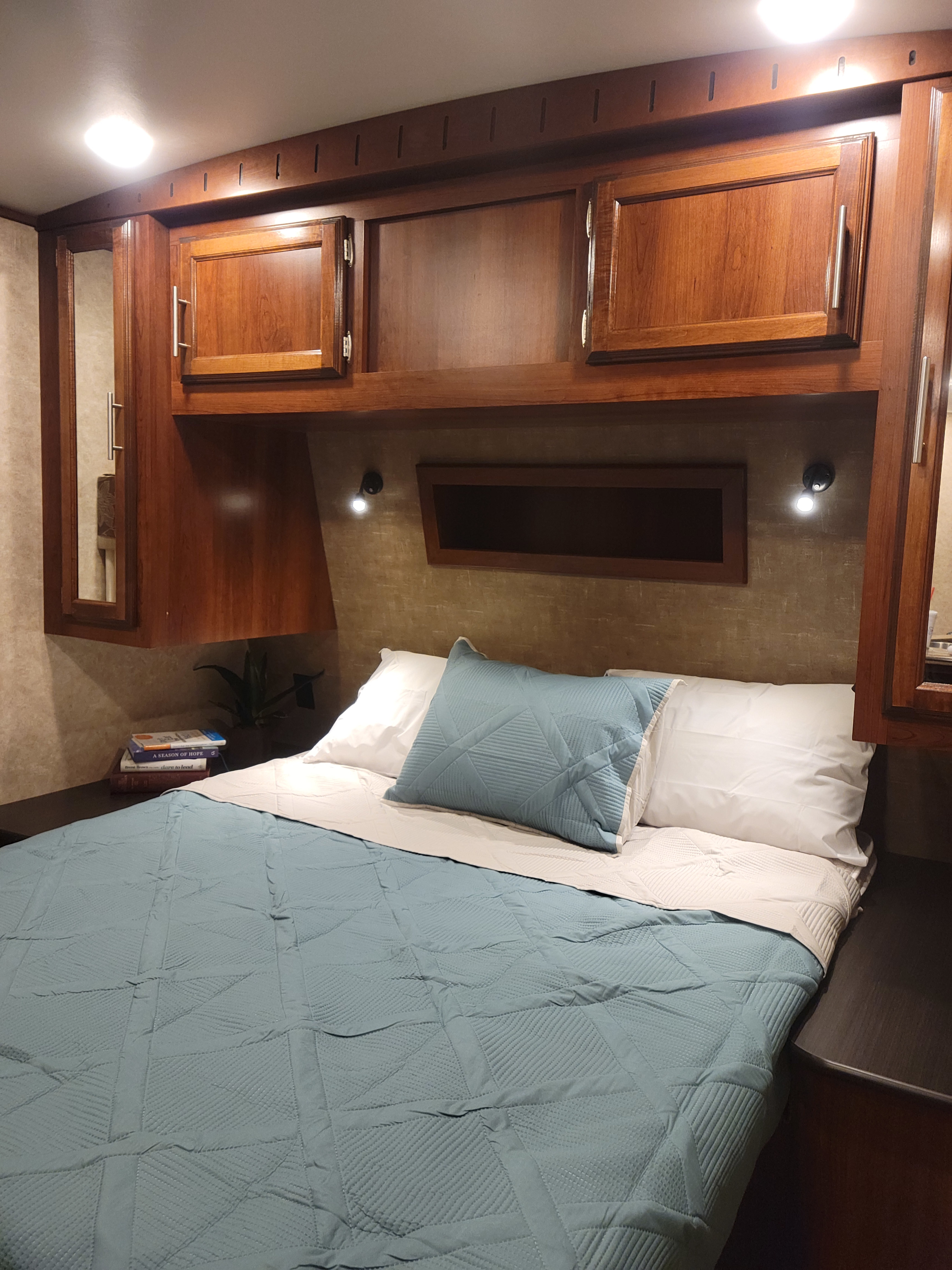 An RV rental bedroom with warm wooden cabinets, a bed with blue and white bedding, and wall-mounted reading lights.. Jayco Eagle 2016