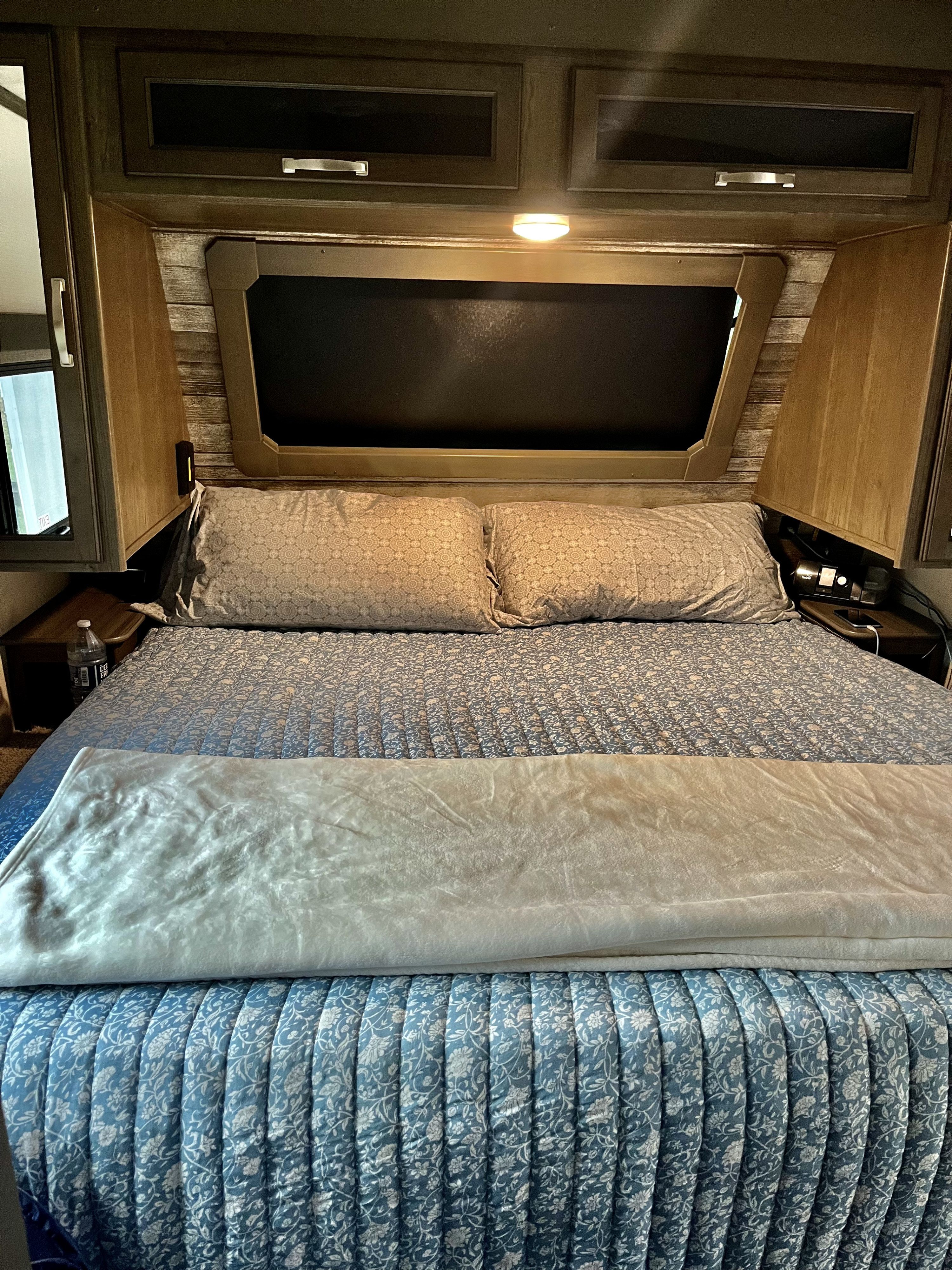 Comfortable RV rental bedroom featuring a blue quilt, light blanket, two pillows, and handy overhead cabinets with a bright light.. Keystone RV Cougar 2022