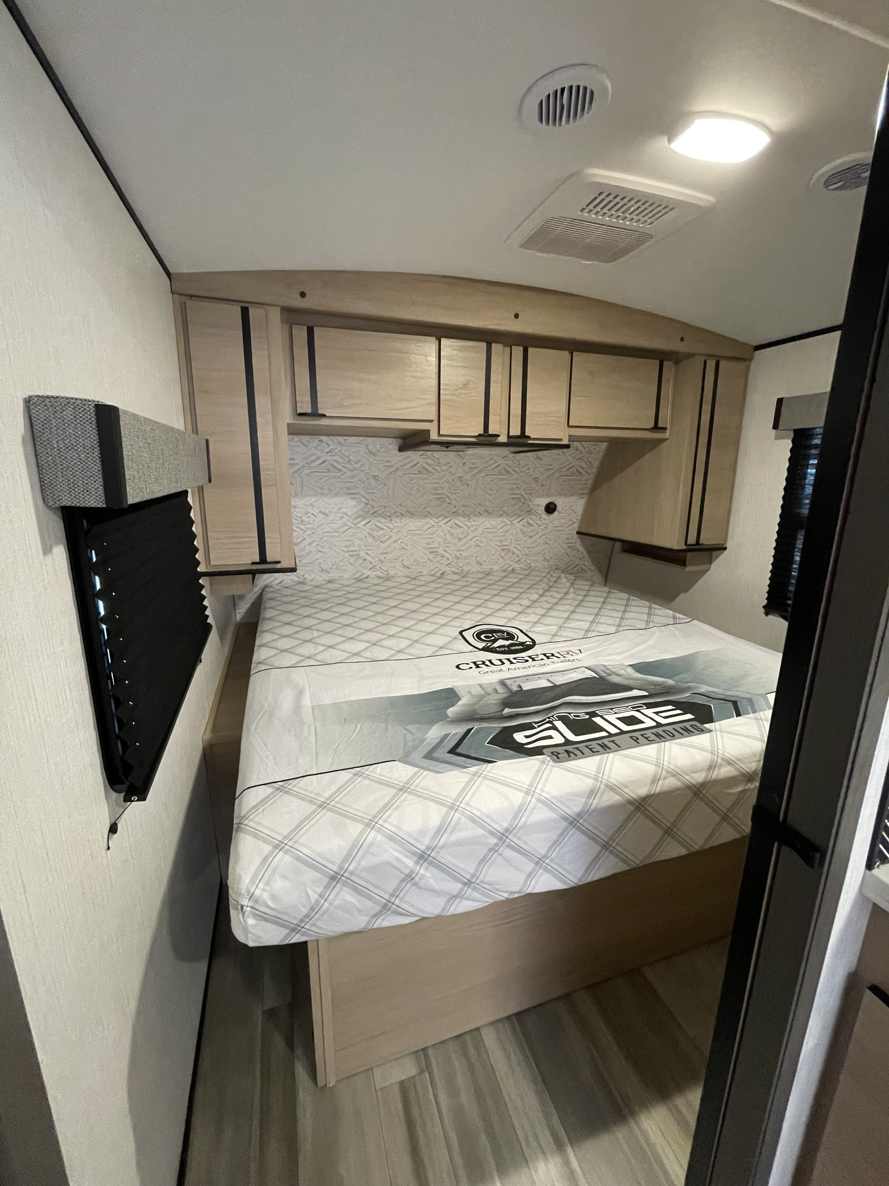 RV rental bedroom featuring a bed, light wood overhead cabinets, and a window with blinds. Clean and compact.. Cruiser Rv Corp Radiance 2022
