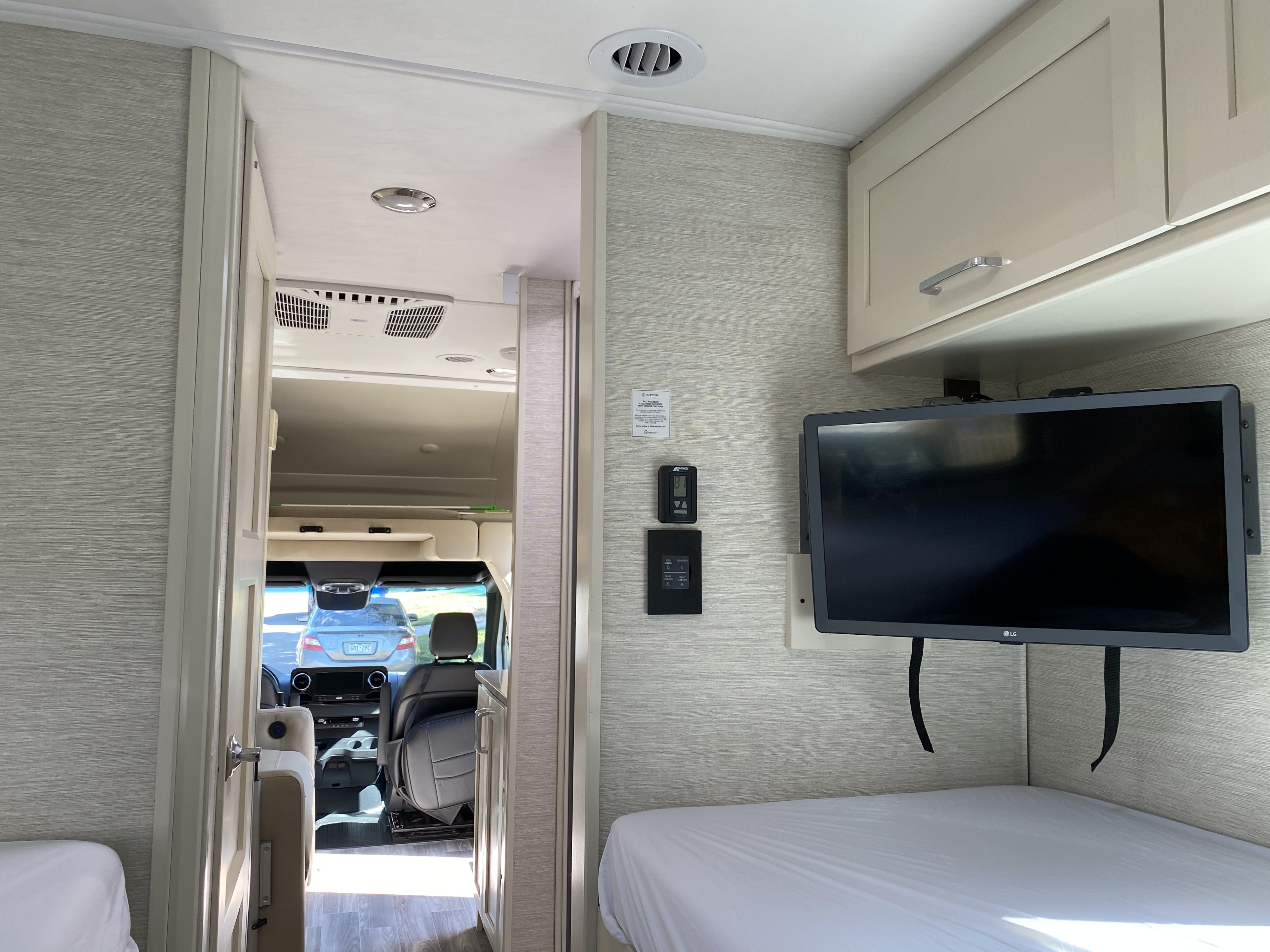 RV rental interior featuring a bedroom with a mounted TV, overhead cabinets, and a doorway leading to the cab area.. Tiffin Motorhomes Wayfarer LW 2023