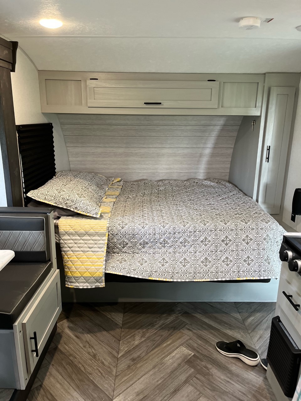 RV rental bedroom with patterned grey/yellow bed, light wood cabinets, and herringbone flooring. Dinette to left.. Forest River Wildwood FSX Plantium 2022