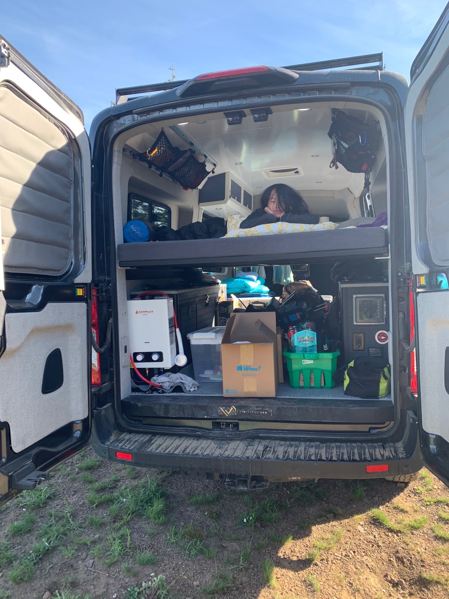 Inside an **RV rental**, a person rests on the top bunk of a camper van. Below, organized storage & portable shower for van life.. Other Transit 2020