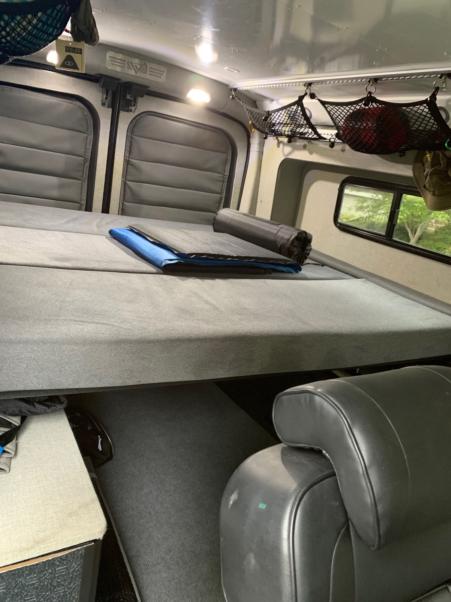 Inside an RV rental: a comfortable bed with a sleeping pad, overhead storage, and a view outside.. Other Transit 2020