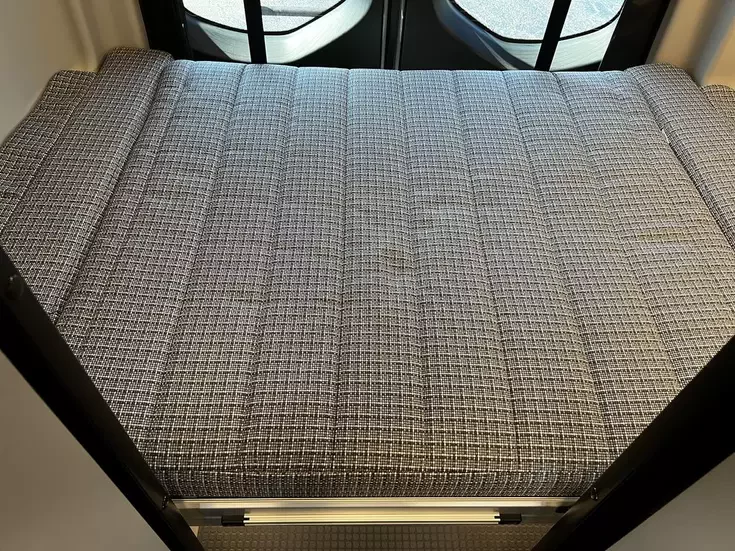 Inside an RV rental: a textured gray RV bed with vertical seams, providing a comfortable sleeping area within the vehicle.. Winnebago Revel 2022