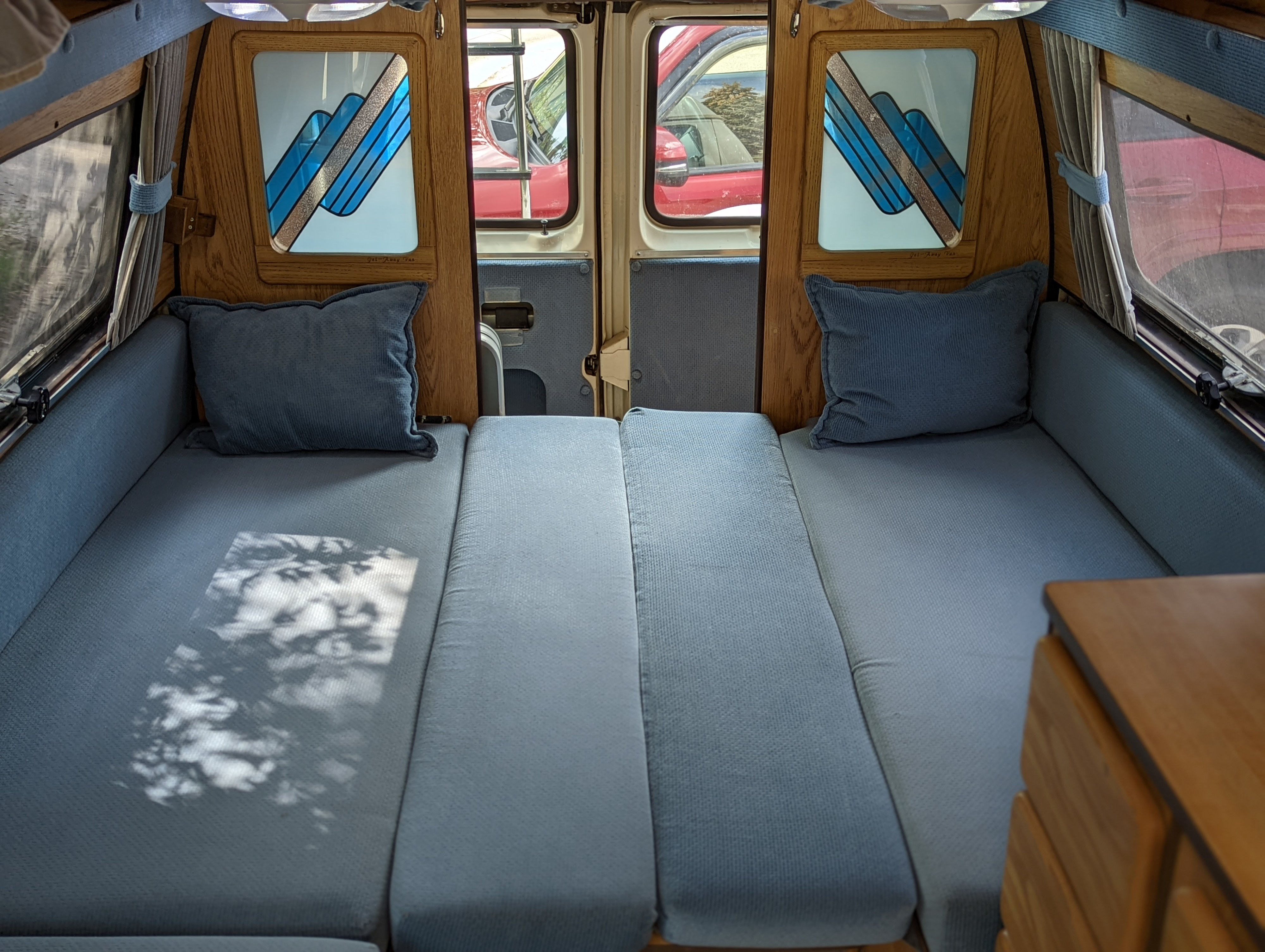 Cozy RV rental interior with blue cushioned beds, pillows, and wooden paneling. Open back doors reveal a red car outside.. Ford Econoline 1988