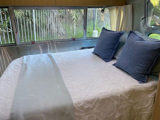 Inside an RV rental: a bright bedroom with white bedding, blue pillows, and large windows revealing tropical greenery.. Airstream Flying Cloud 2015