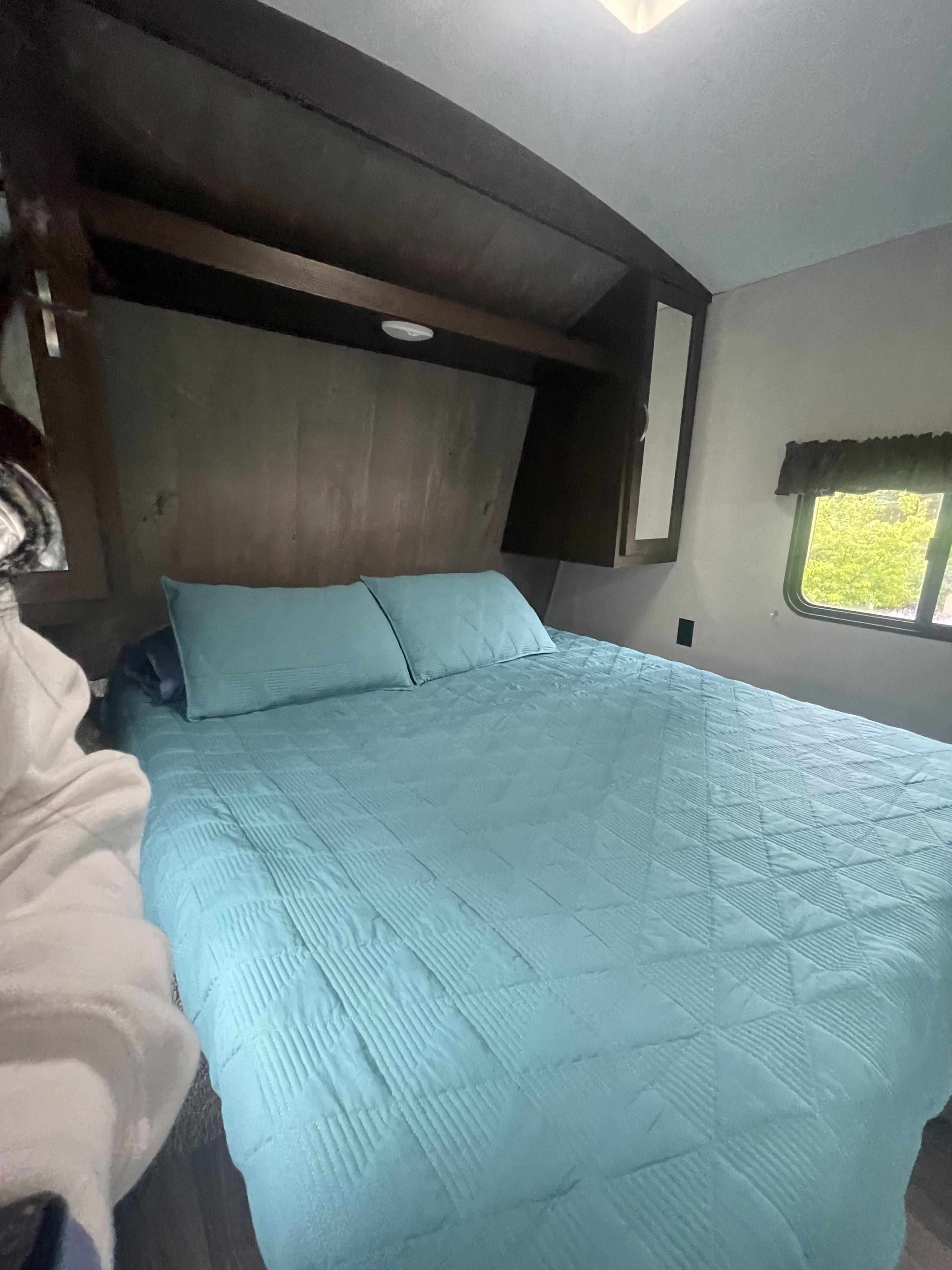 Inside a comfortable RV rental bedroom, featuring a blue bed with pillows, overhead cabinets, and a window view.. Keystone RV Springdale 2020