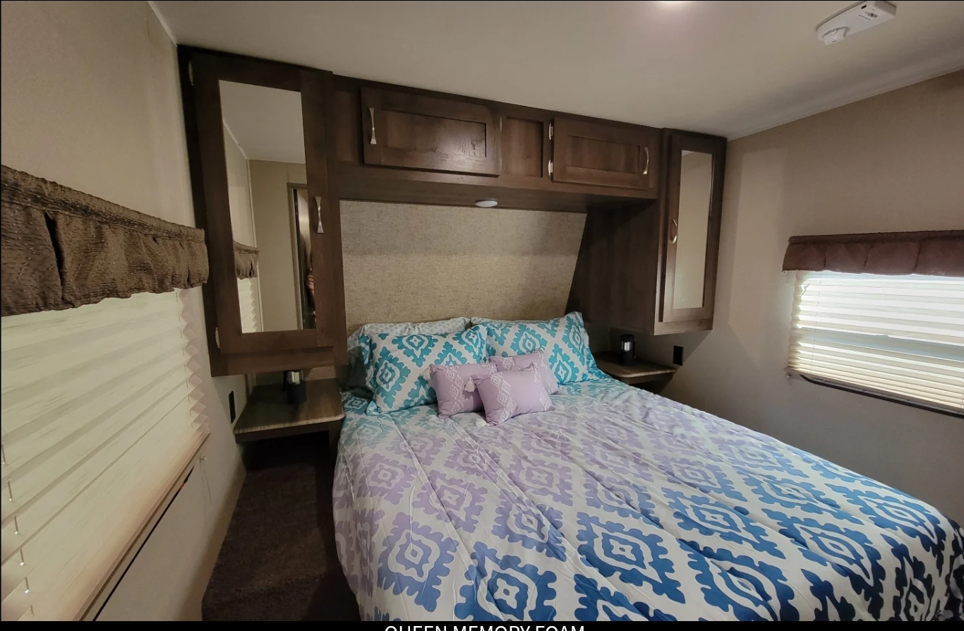 Cozy RV rental bedroom features a queen bed with blue/purple bedding, overhead cabinets, and window blinds.. Forest River Palomino Puma 2017