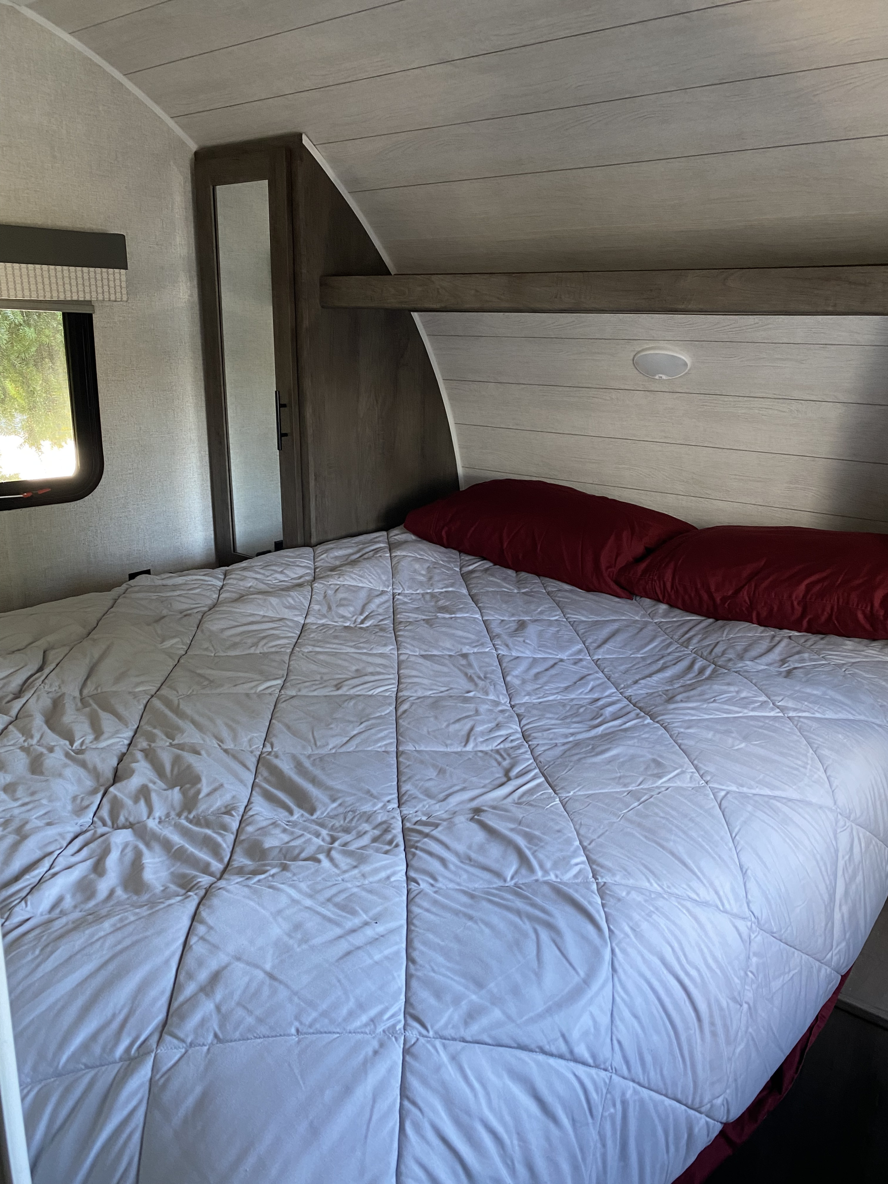 Inside an RV rental, a neatly made bedroom featuring a comfy bed, wood-paneled ceiling/headboard, and a window.. Forest River Wildwood X-Lite 2021