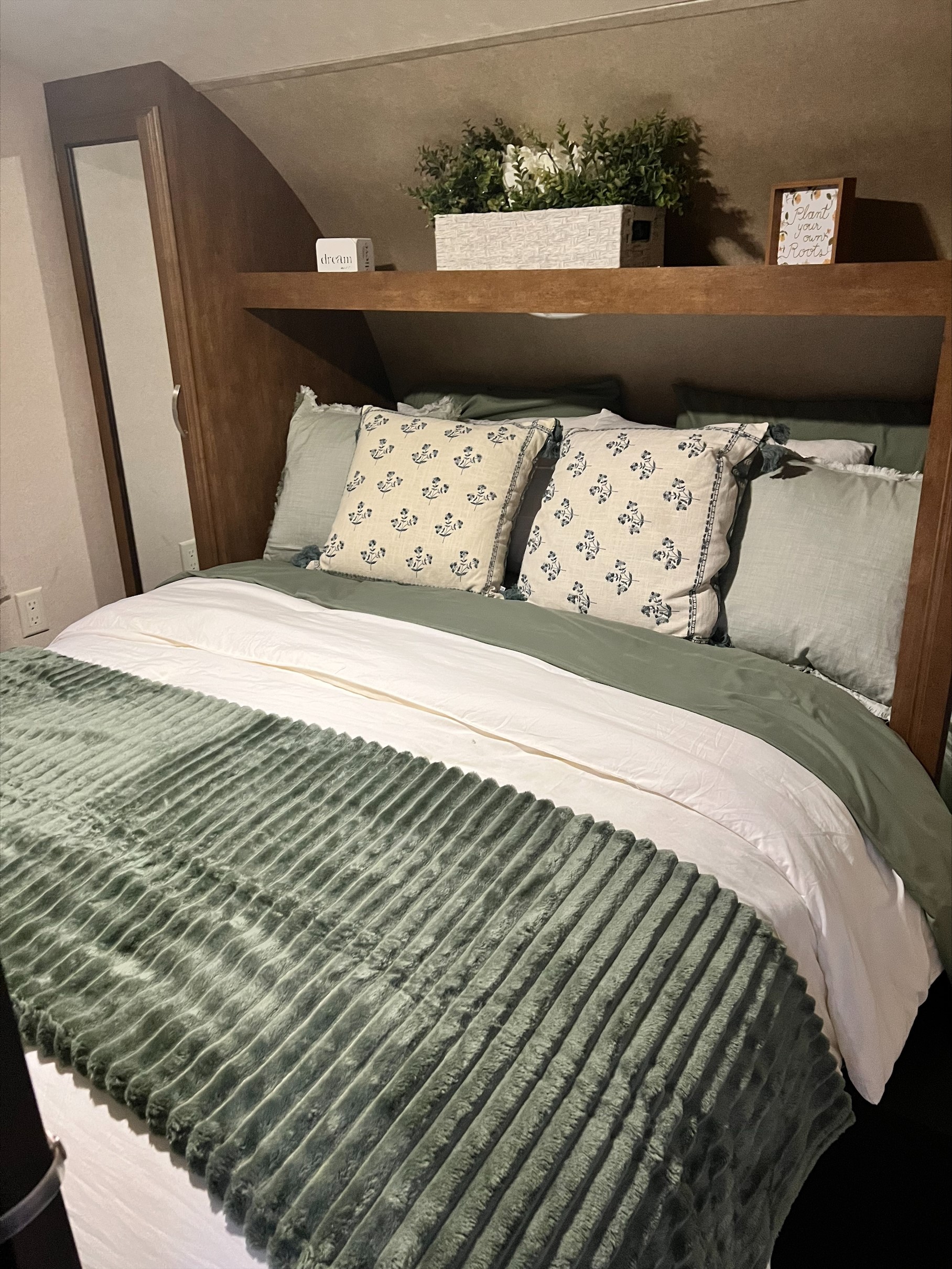 This RV rental bedroom offers a snug retreat with green & white bedding, floral pillows, and charming shelf decor.. Forest River Wildwood 2017