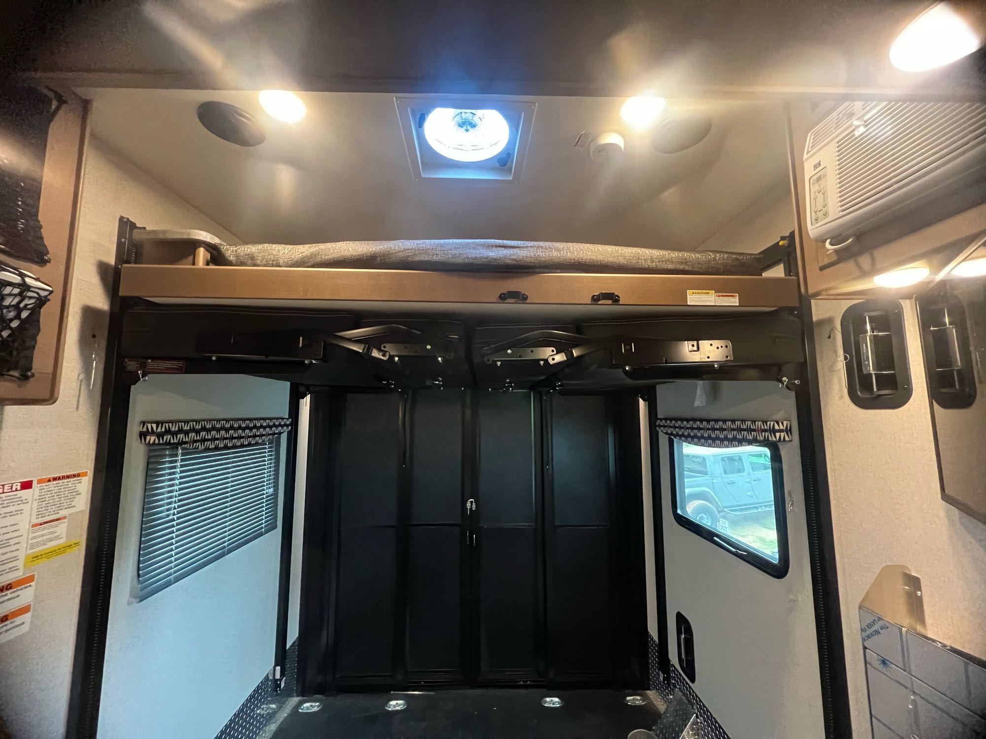 Inside an RV rental: Rear view of a toy hauler's interior with a raised bunk, ceiling lights, A/C, and large cargo door.. Thor Outlaw 2022