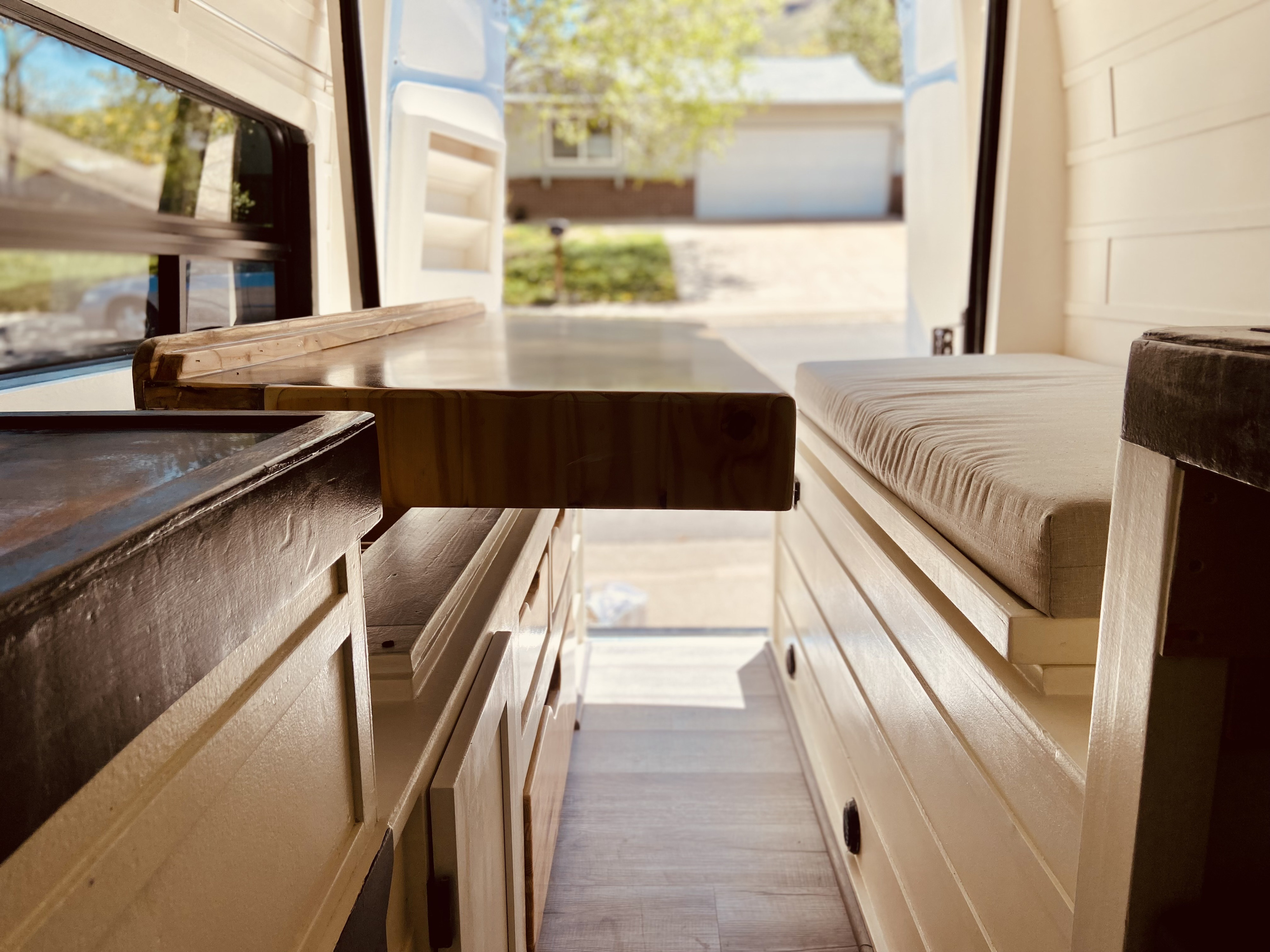 View from inside an RV rental: custom wood counter, padded bench, and light cabinetry. Open doors reveal a street.. Mercedes Sprinter 2021