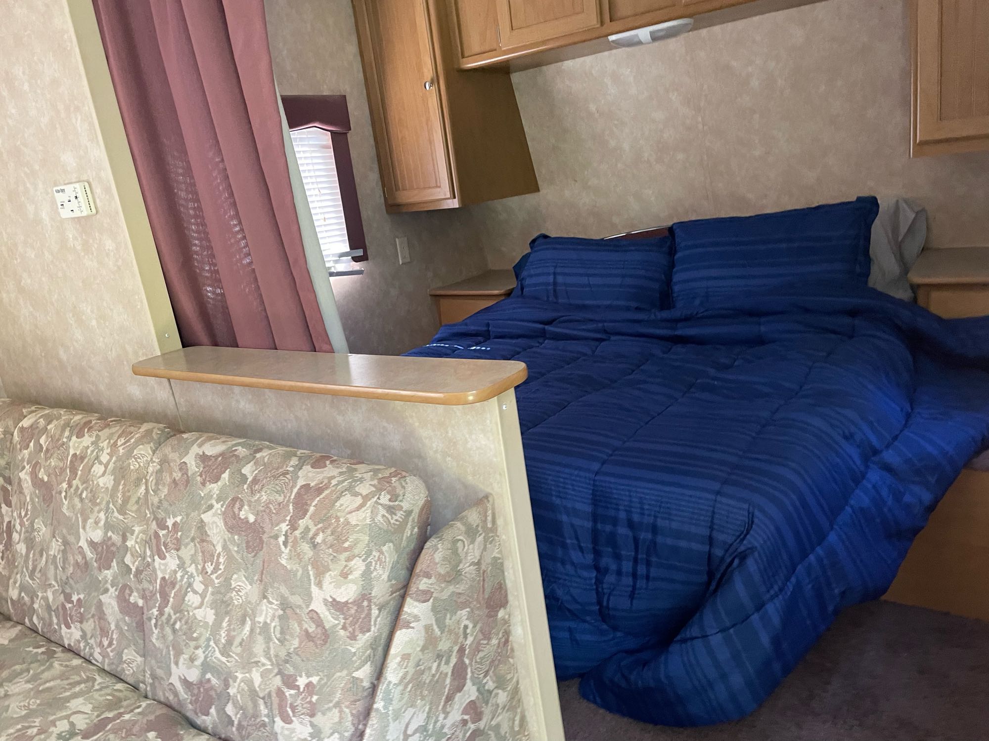Cozy RV rental interior showing a compact bedroom with blue bedding, overhead cabinets, and an adjacent living area sofa.. Dutchmen Sport 2004