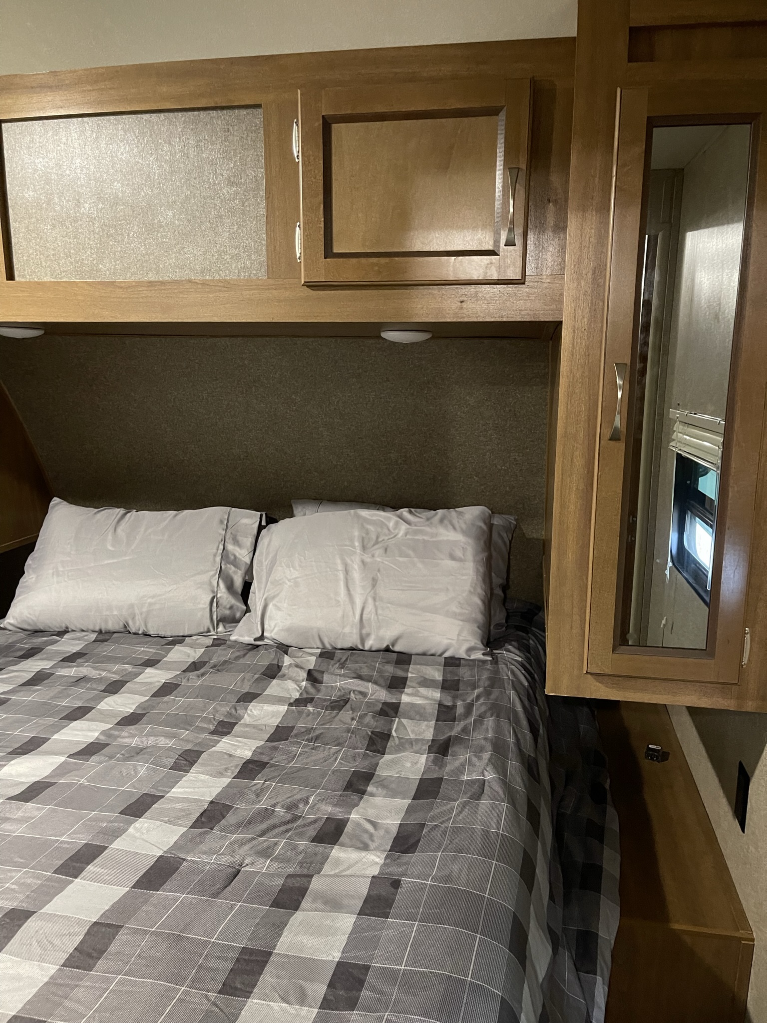 Cozy RV rental bedroom interior features a plaid bed, overhead wood cabinets, and a mirrored closet door. Ideal for travel.. Coachmen Catalina 2019