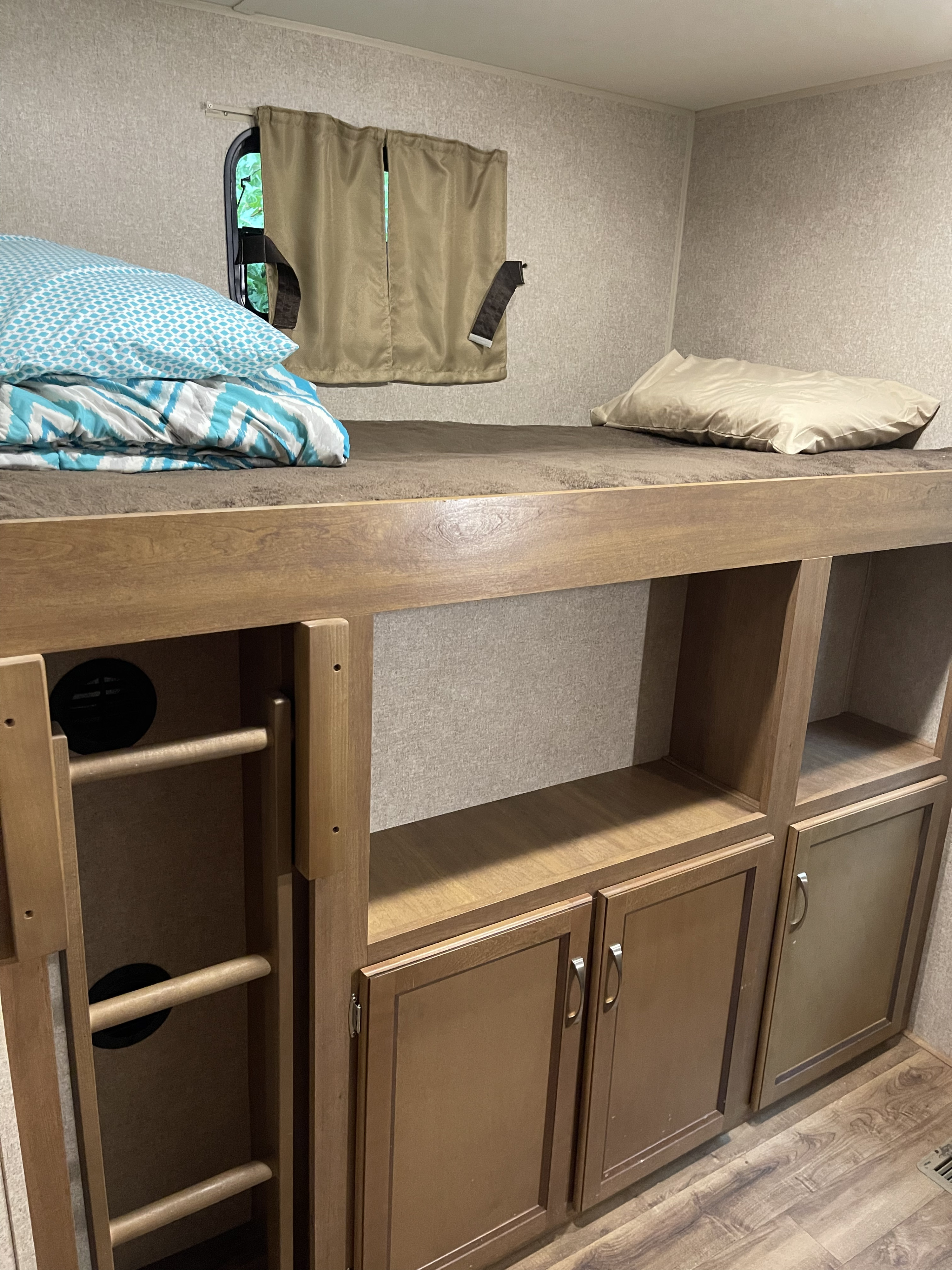Inside an RV rental: a cozy bunk bed with blue bedding, a ladder, and built-in wood cabinets for storage. Window with curtains visible.. Coachmen Catalina 2019