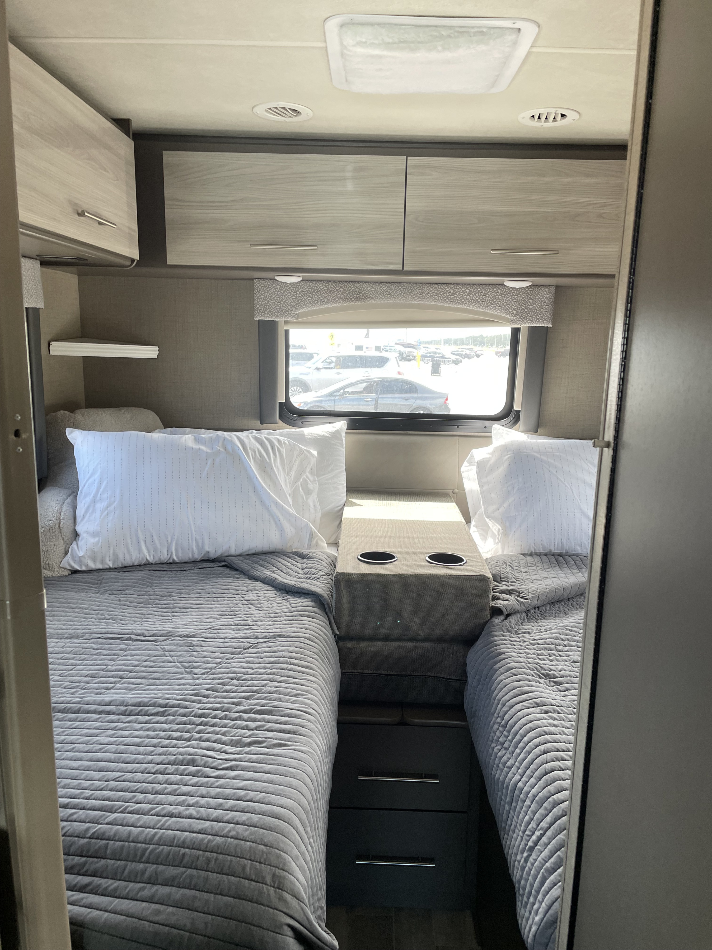 RV rental bedroom: twin beds with grey quilts, central console with cupholders, overhead storage, and bright window view.. Thor Vegas 2020