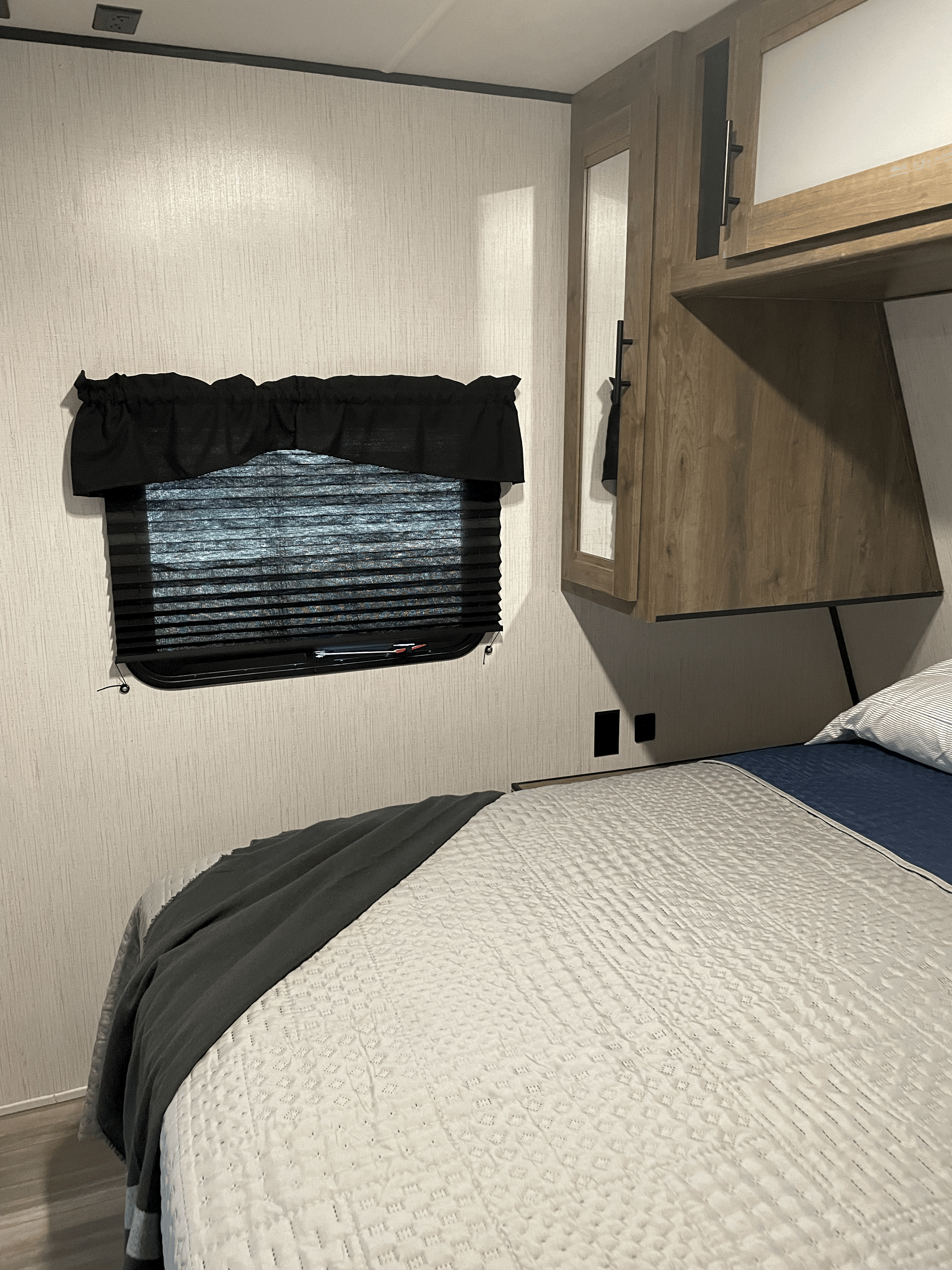 This RV rental bedroom features a comfy bed with a quilted cover, dark throw, window blinds, and overhead storage cabinets.. Heartland RVs Pioneer 2021