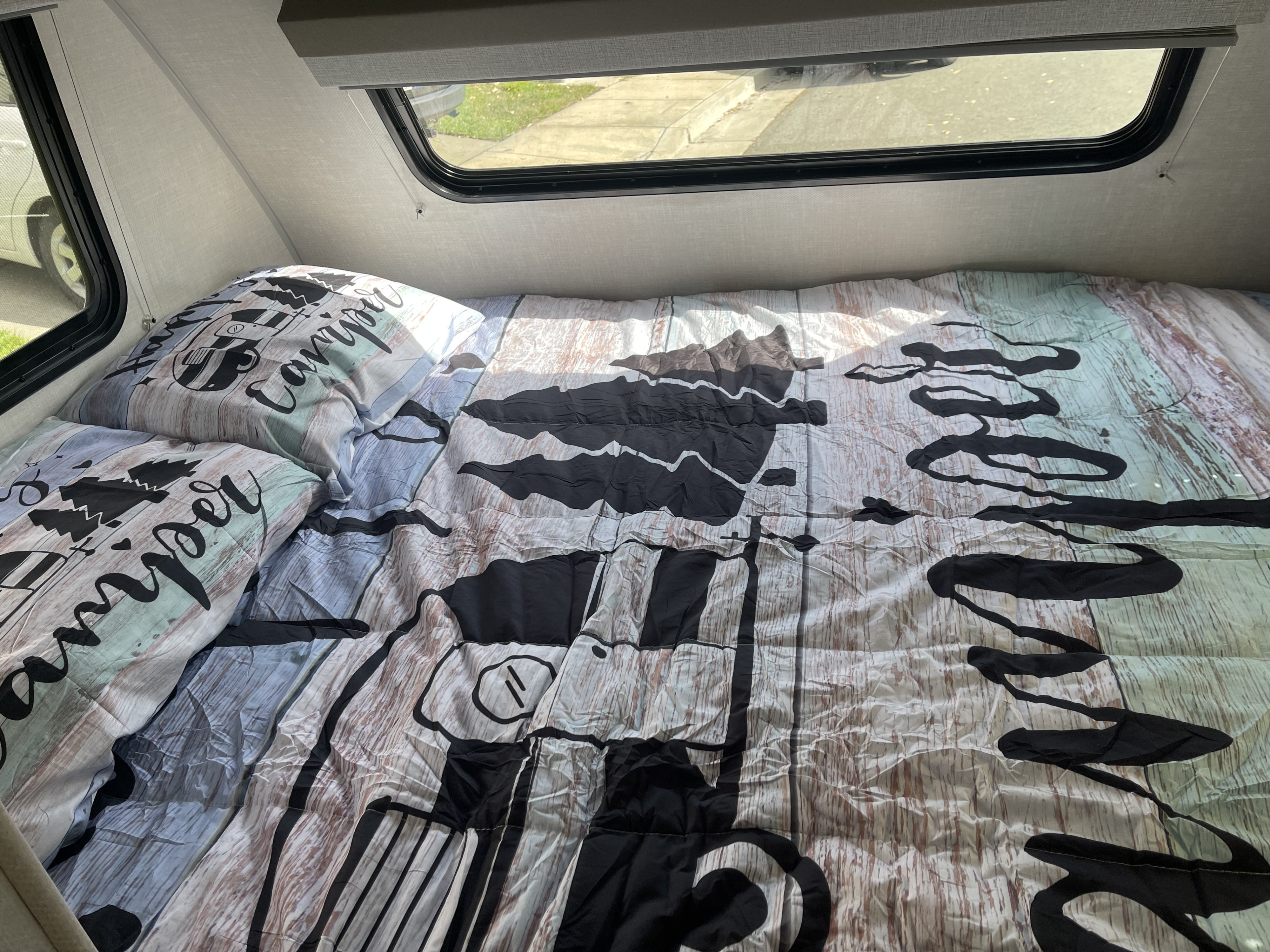 Cozy RV rental bed featuring "Happy Camper" themed bedding, with sunlight streaming in through the window, revealing outdoor scenery.. Forest River R-Pod 171 2023