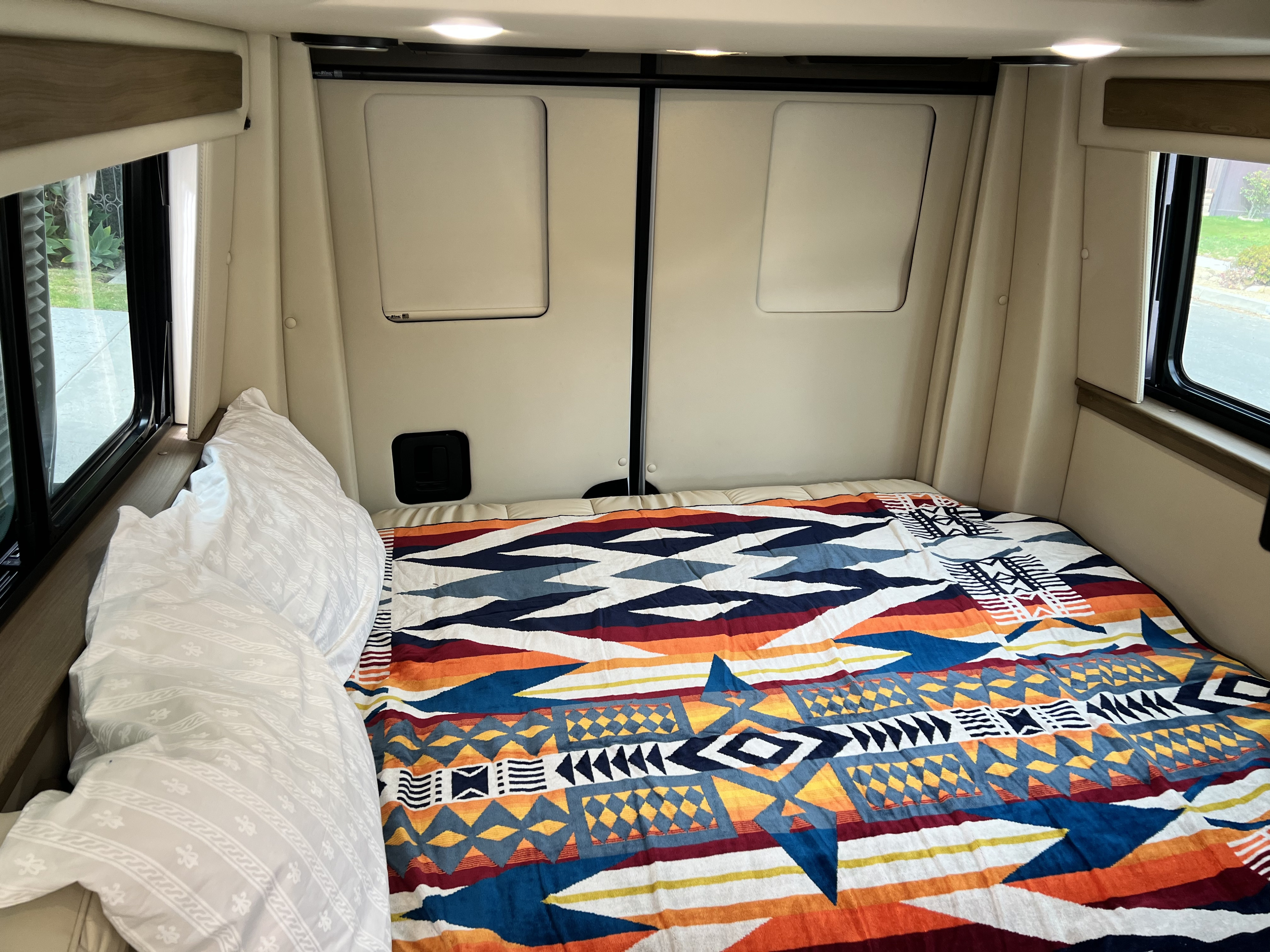 Cozy RV rental bedroom with a colorful geometric blanket, white pillows, and natural light from side windows.. Midwest Automotive Designs Midwest Automotive Designs Class B 2021