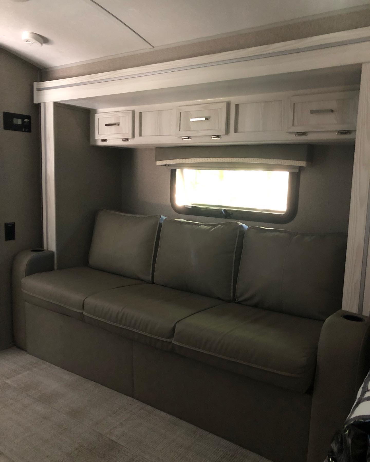Modern RV rental living area featuring a gray sofa with cupholder, overhead cabinets, and a bright window.. Forest River Rockwood Geo Pro 2020