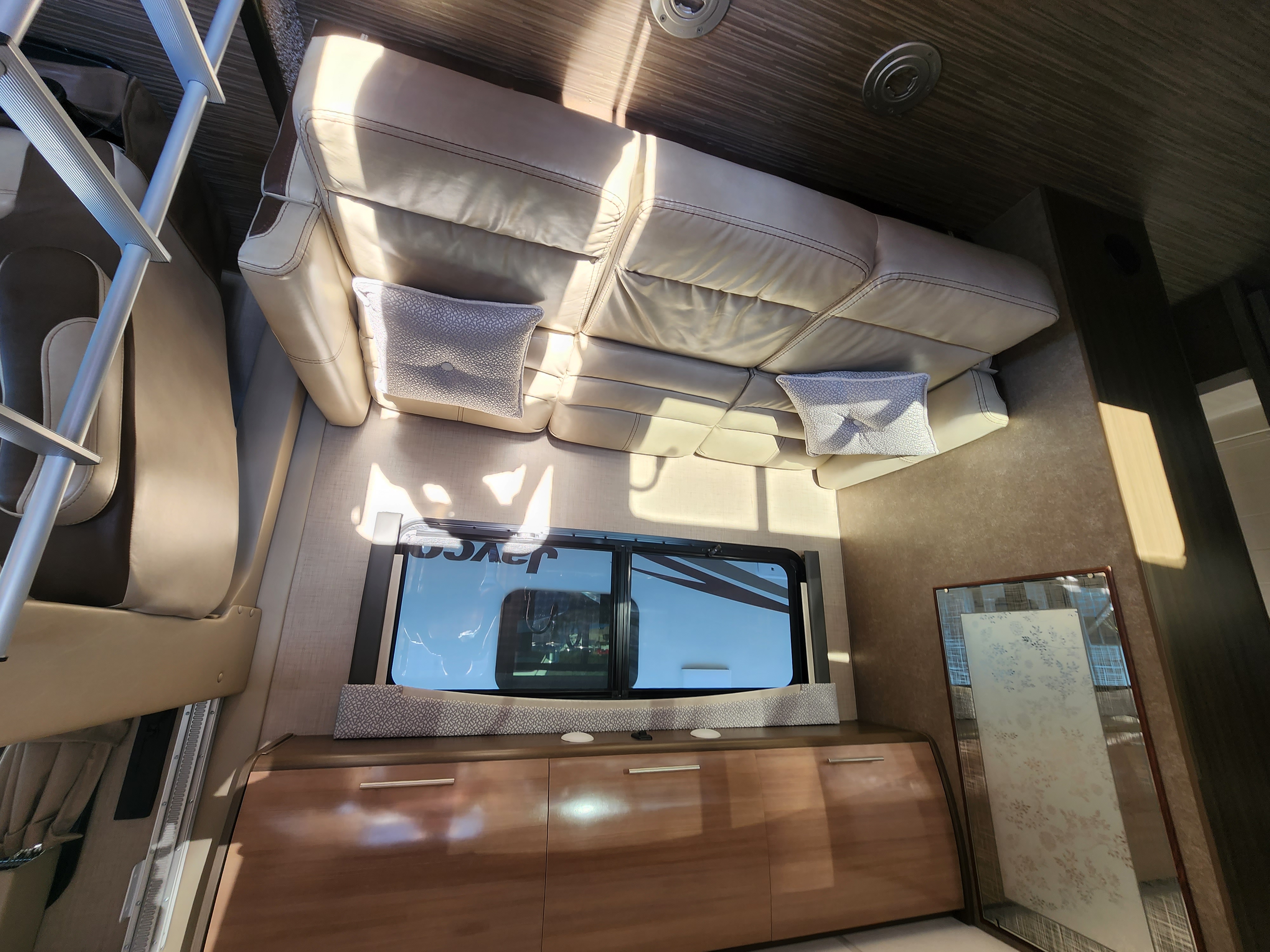 Bright RV rental interior: cream leather sofa, entertainment unit, & window revealing the Jayco logo. Ladder on left.. Thor Vegas 2019