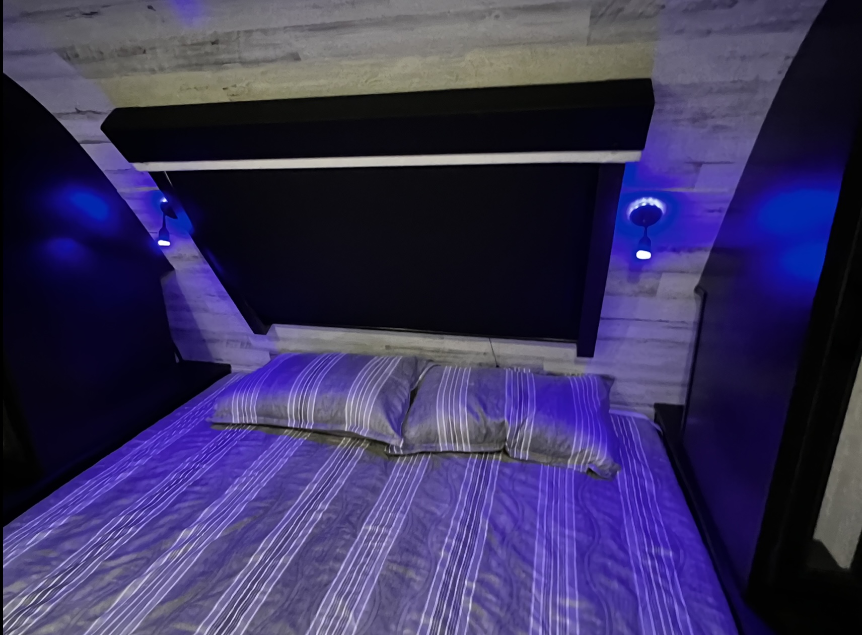 Modern RV rental bedroom with cozy blue lighting, striped bedding, and a sleek headboard, offering a comfortable travel space.. Jayco White Hawk 2022