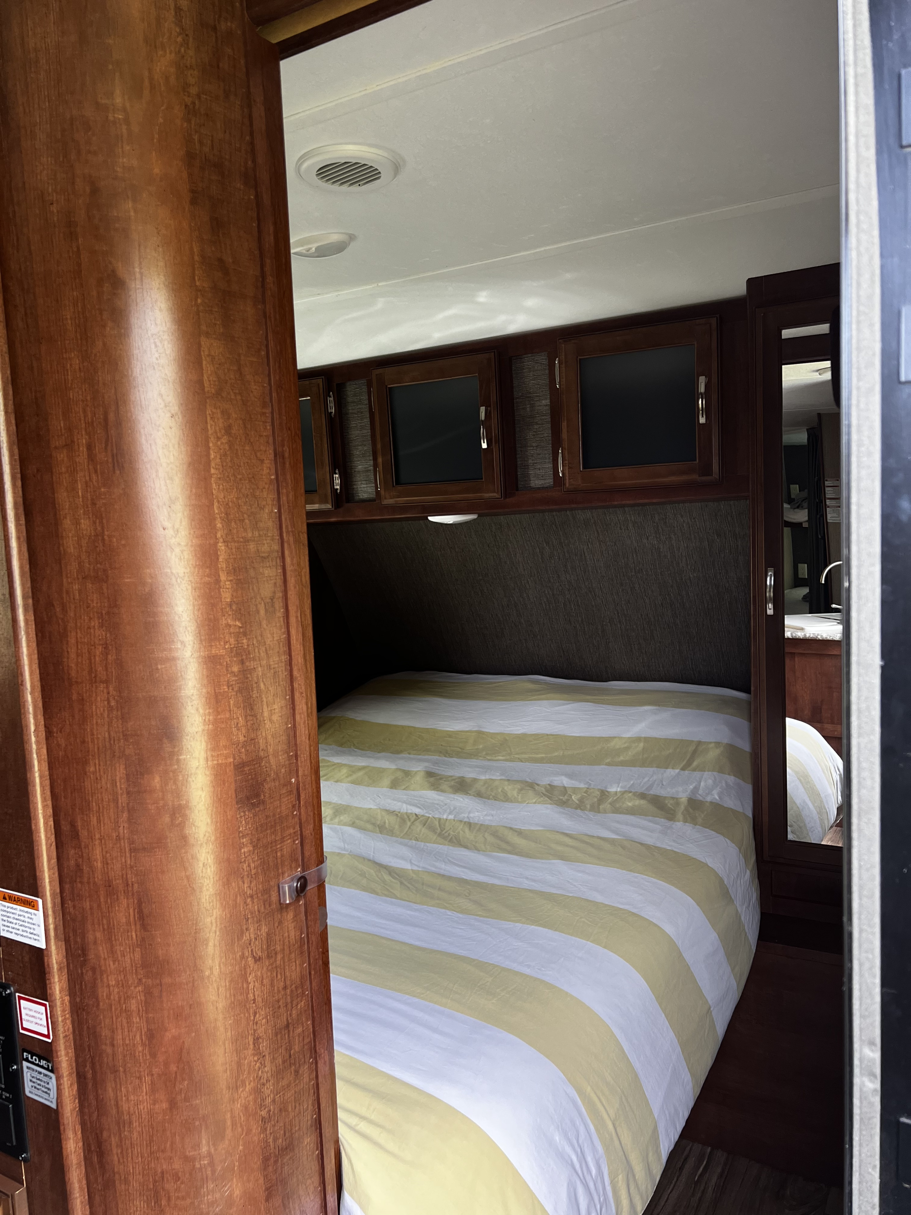 RV rental bedroom showcasing a yellow/white striped bed, rich wooden cabinetry overhead, and a mirror in a compact space.. Keystone RV Passport Grand Touring 2016