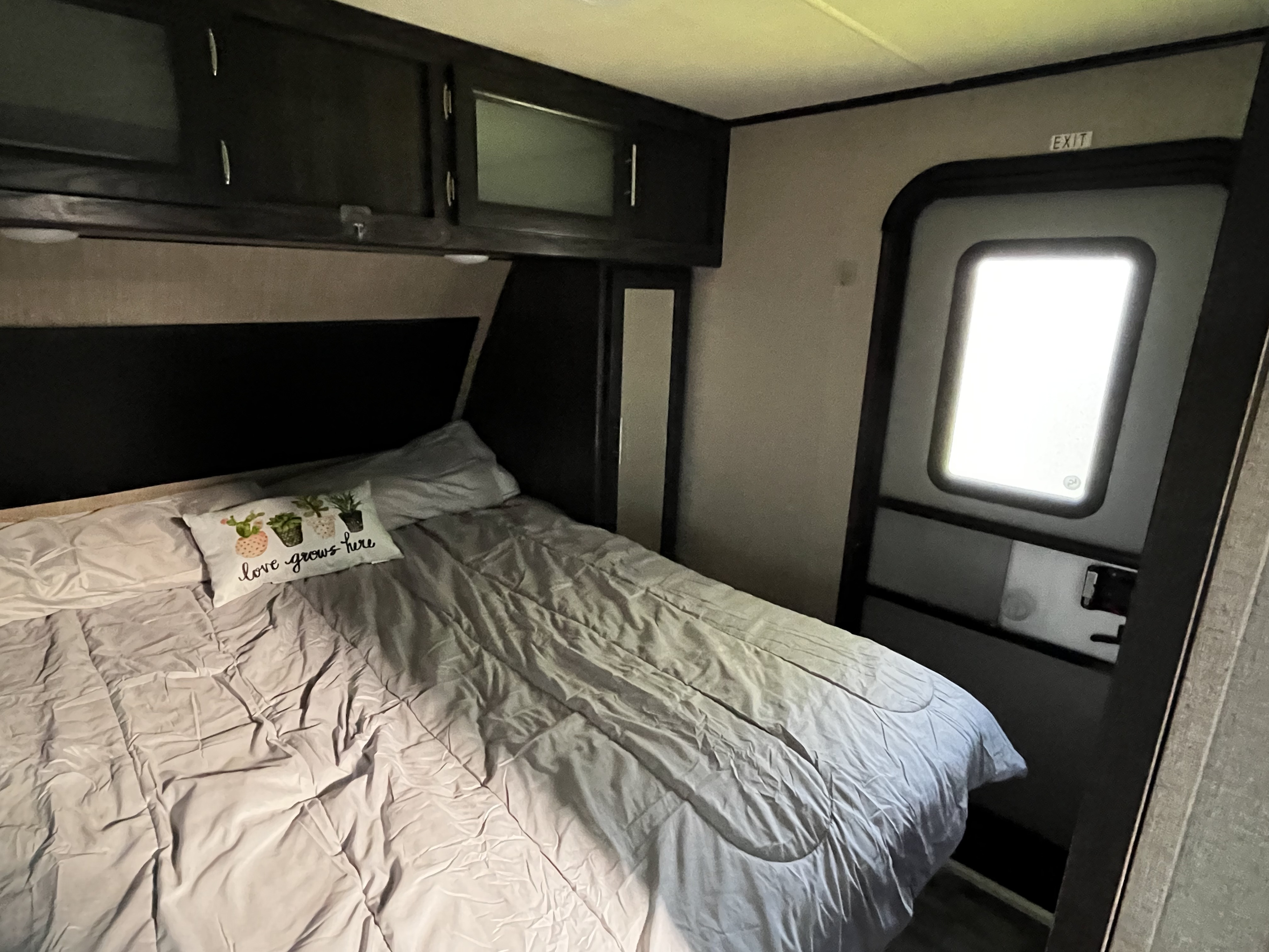Cozy RV rental bedroom featuring a bed with a "love grows here" pillow, overhead cabinets, and an exit door.. Dutchmen Kodiak Ultra Lite 2019