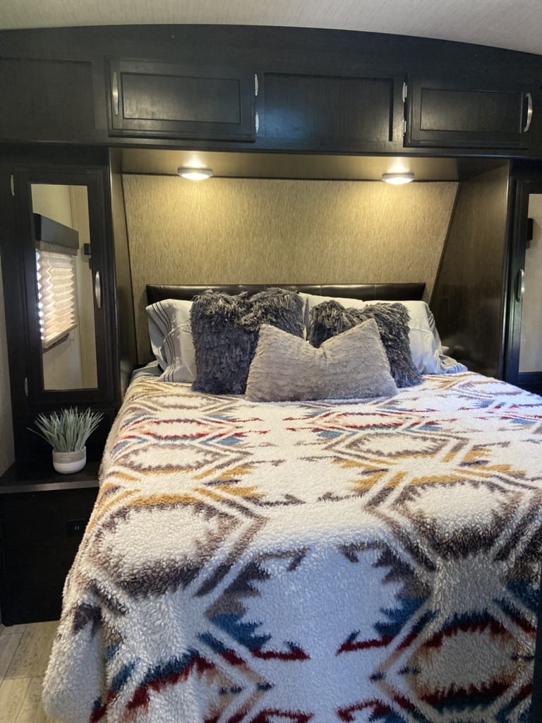 Cozy RV rental bedroom features a bed with a patterned blanket, overhead storage, and soft lighting for a comfortable stay.. Grand Design Imagine 2017