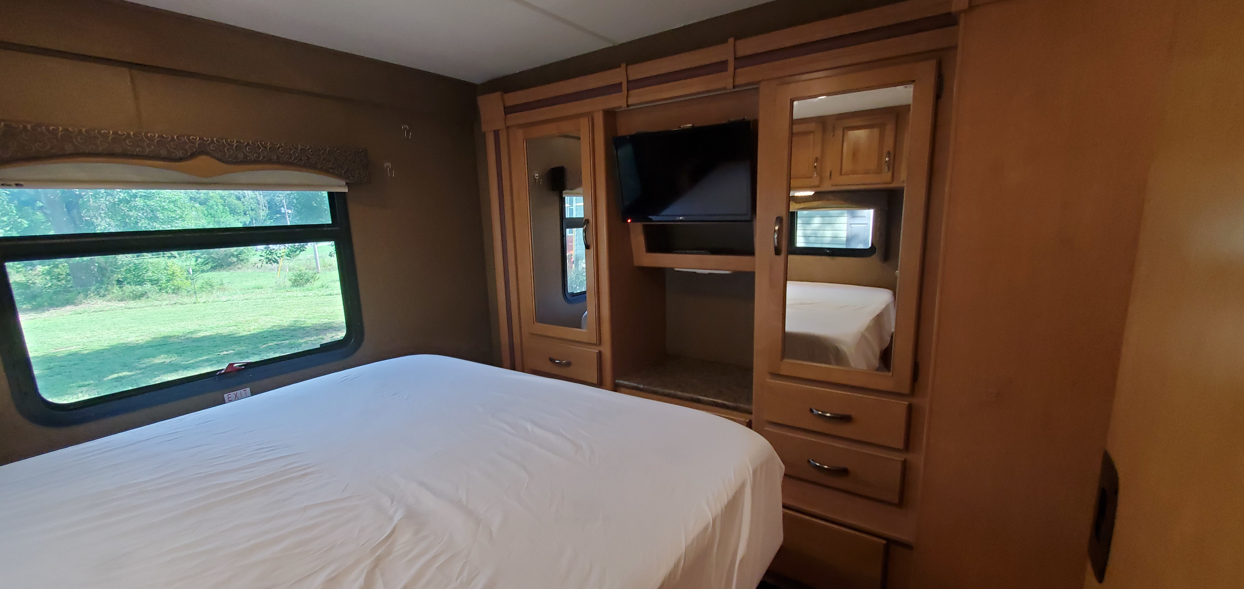 Spacious RV rental bedroom featuring a comfy white bed, large window with green outdoor view, TV, and wood storage.. Thor Four Winds 31E 2015