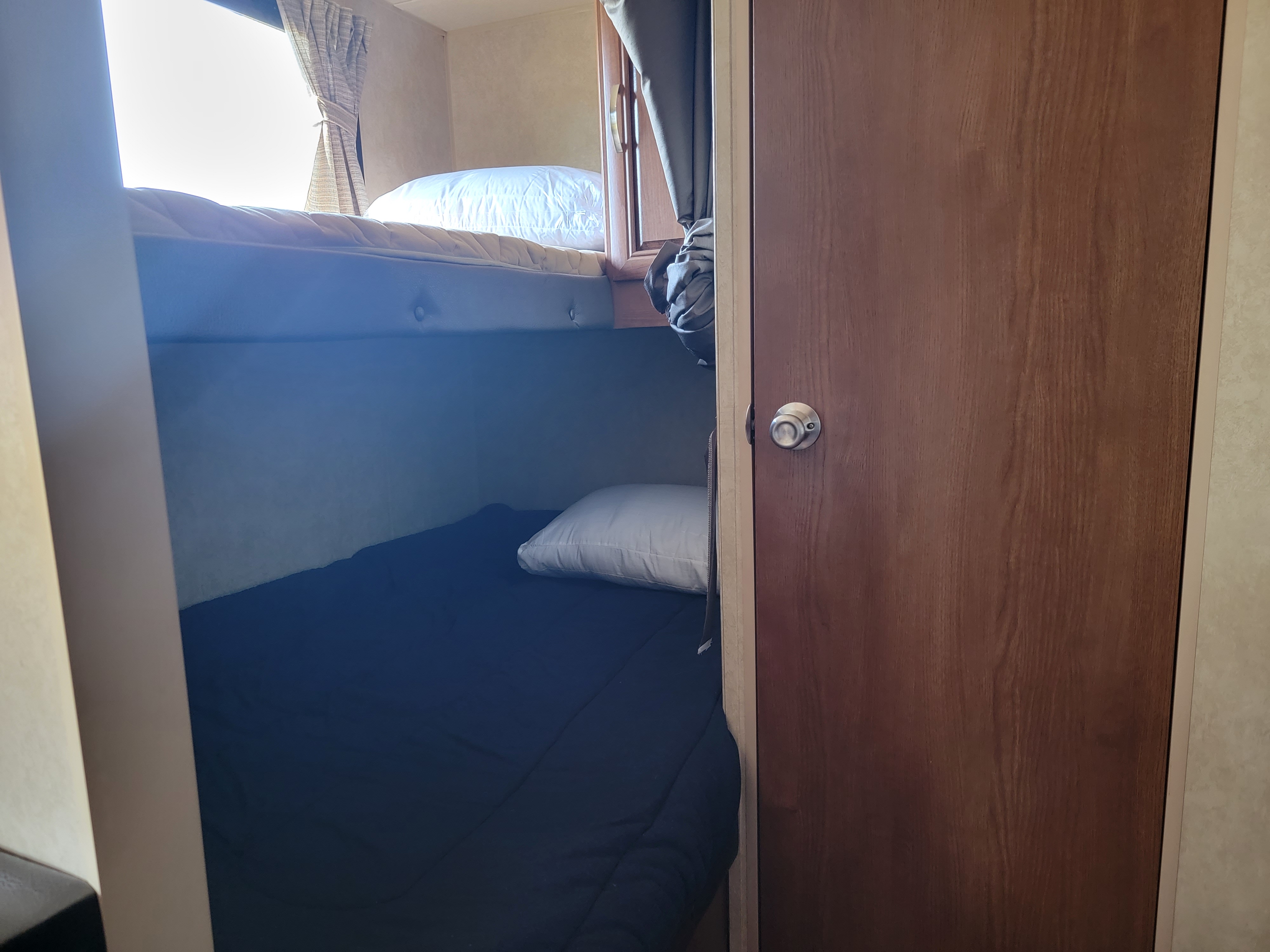 Inside an **rv rental**, showing bunk beds with a pillow on the lower bed, a bright window with curtains, and a wooden door.. Forest River Salem Cruise Lite 2014