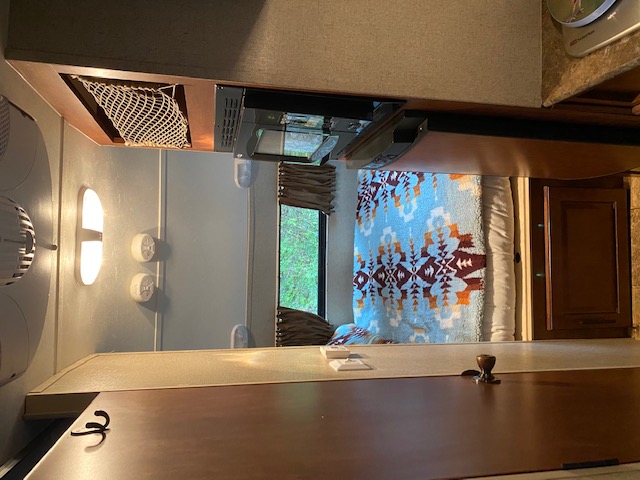 This RV rental's interior features a kitchen counter, microwave, and a cozy bed with a blue patterned blanket, looking out a window.. Forest River Hood River R Pod 2013