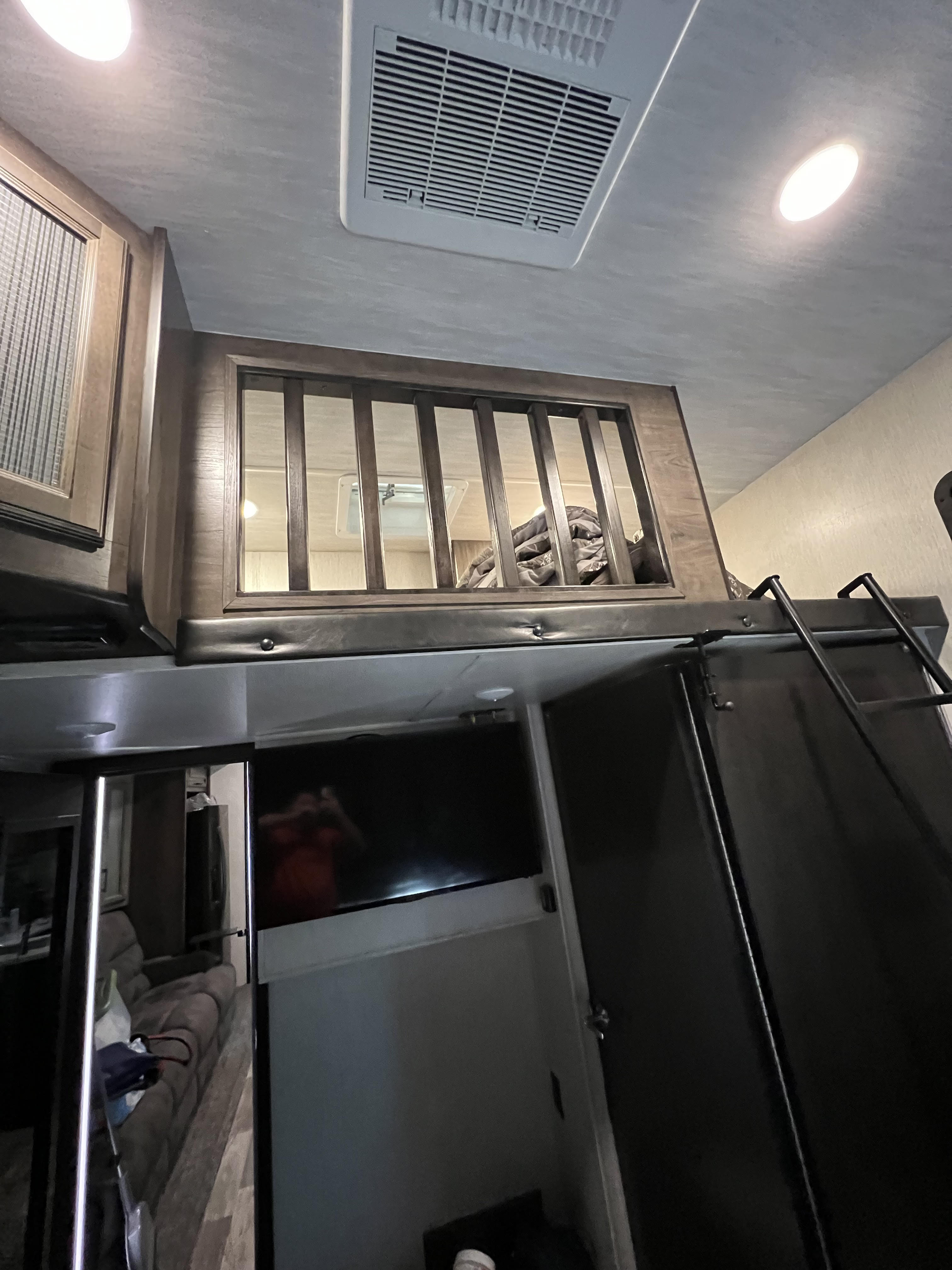 RV rental interior: View up at a wooden loft bed with railing, ladder, AC, lights, and a TV mounted below.. Heartland Cyclone 2019