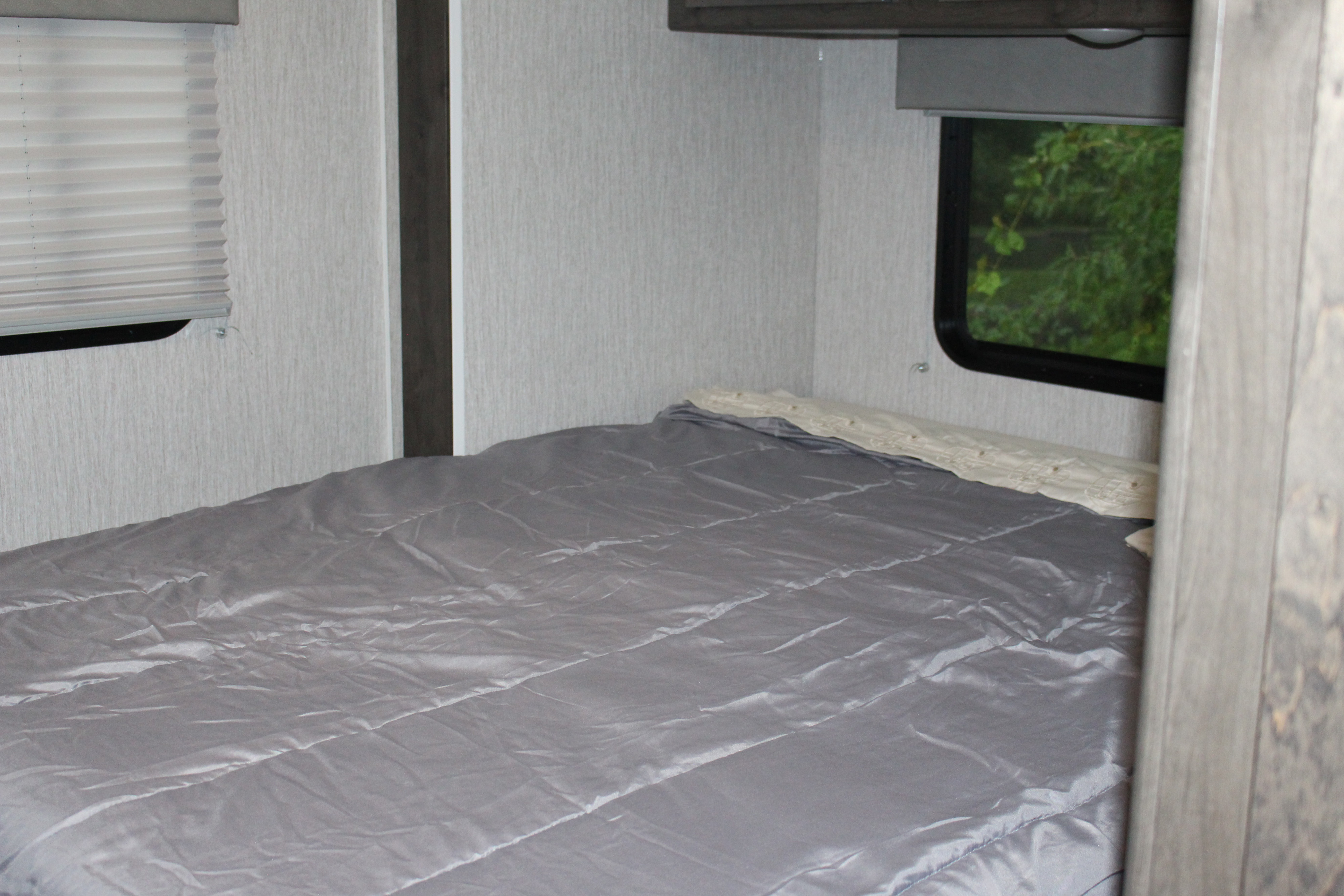 Interior of an RV rental bedroom showing a gray bed, light walls, and windows with blinds and outdoor greenery.. Forest River Coachmen Leprechaun 2022