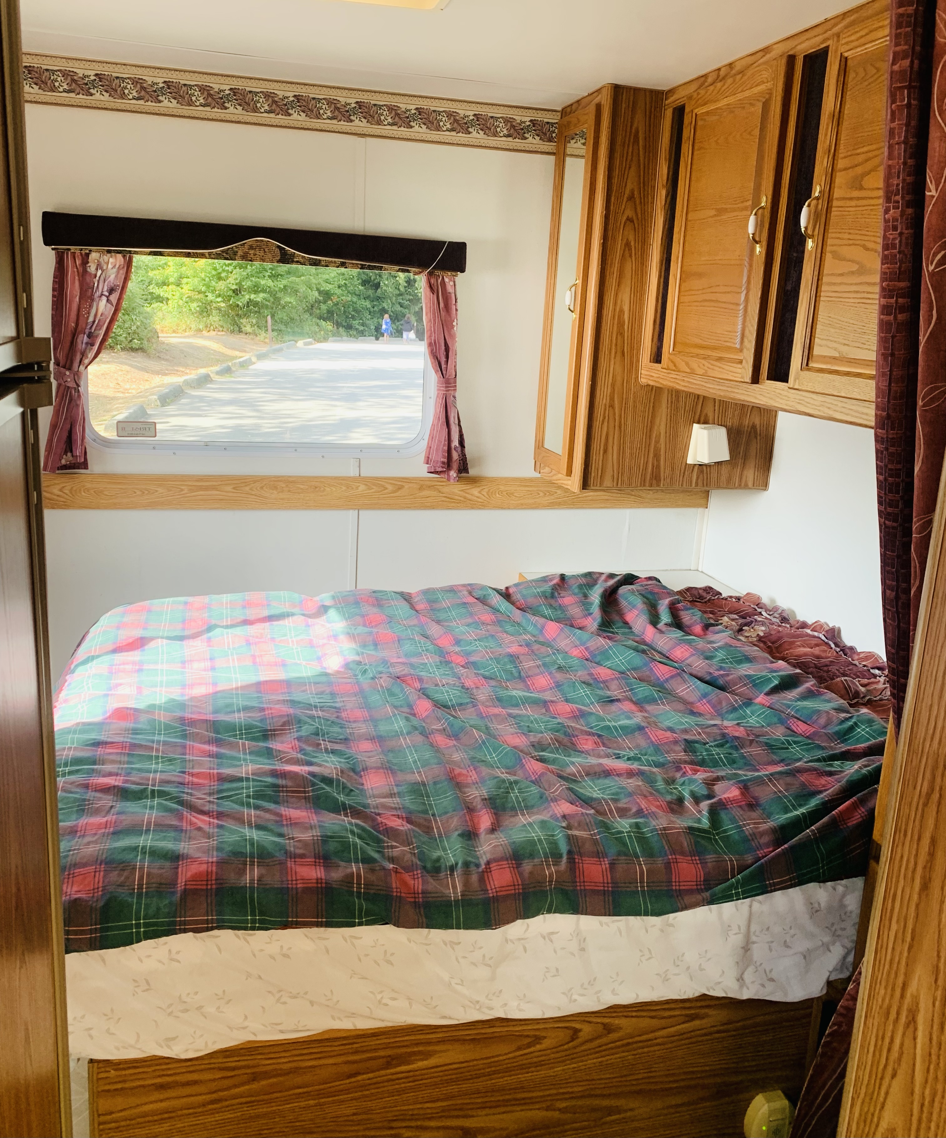 Cozy RV rental bedroom with a plaid bed, wooden cabinets, and sunlit window view of a road and trees.. Slumber Queen Adventurer 2001