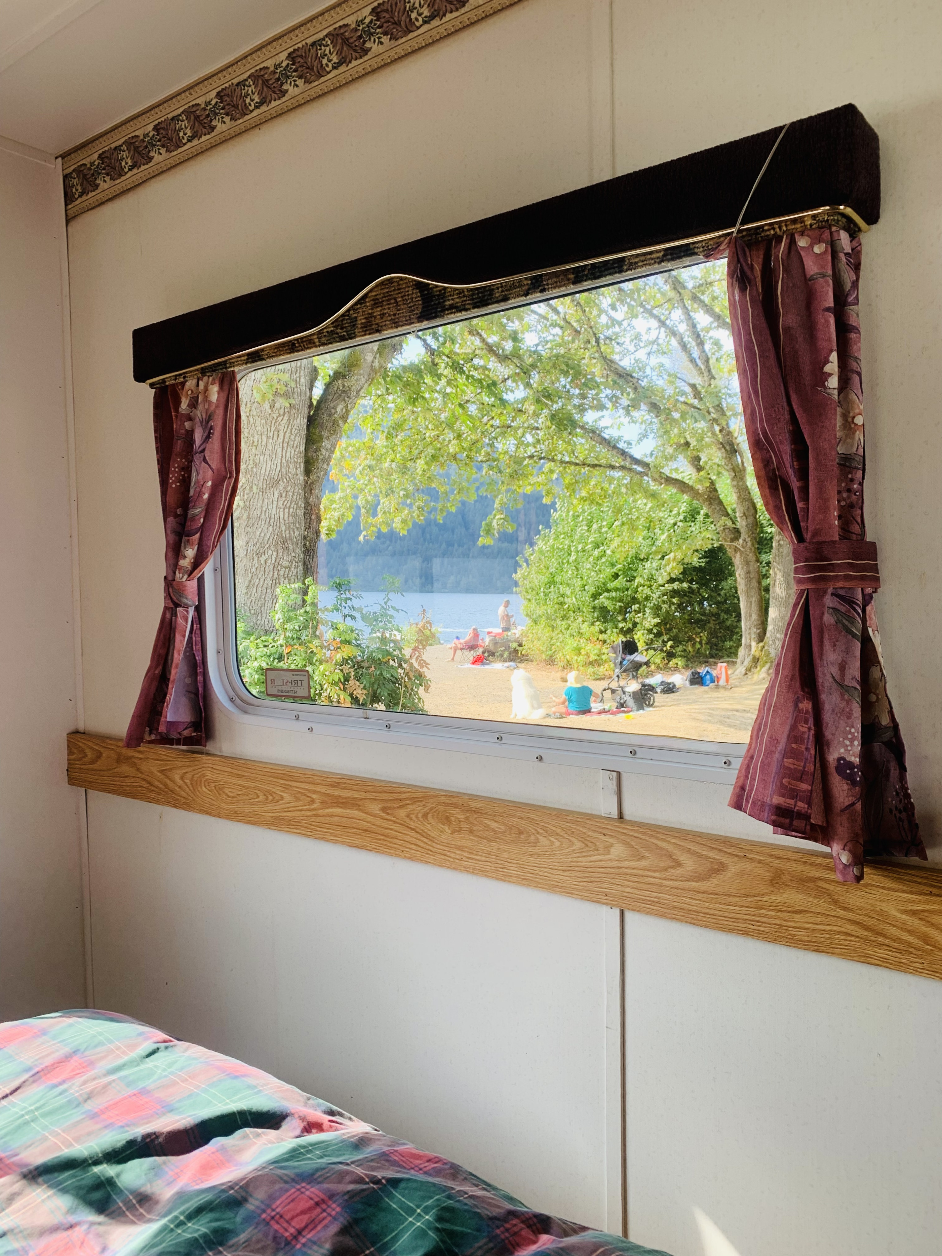 Relaxing RV rental interior showing a plaid bed and a window view of a lakeside camping spot with people and a dog.. Slumber Queen Adventurer 2001