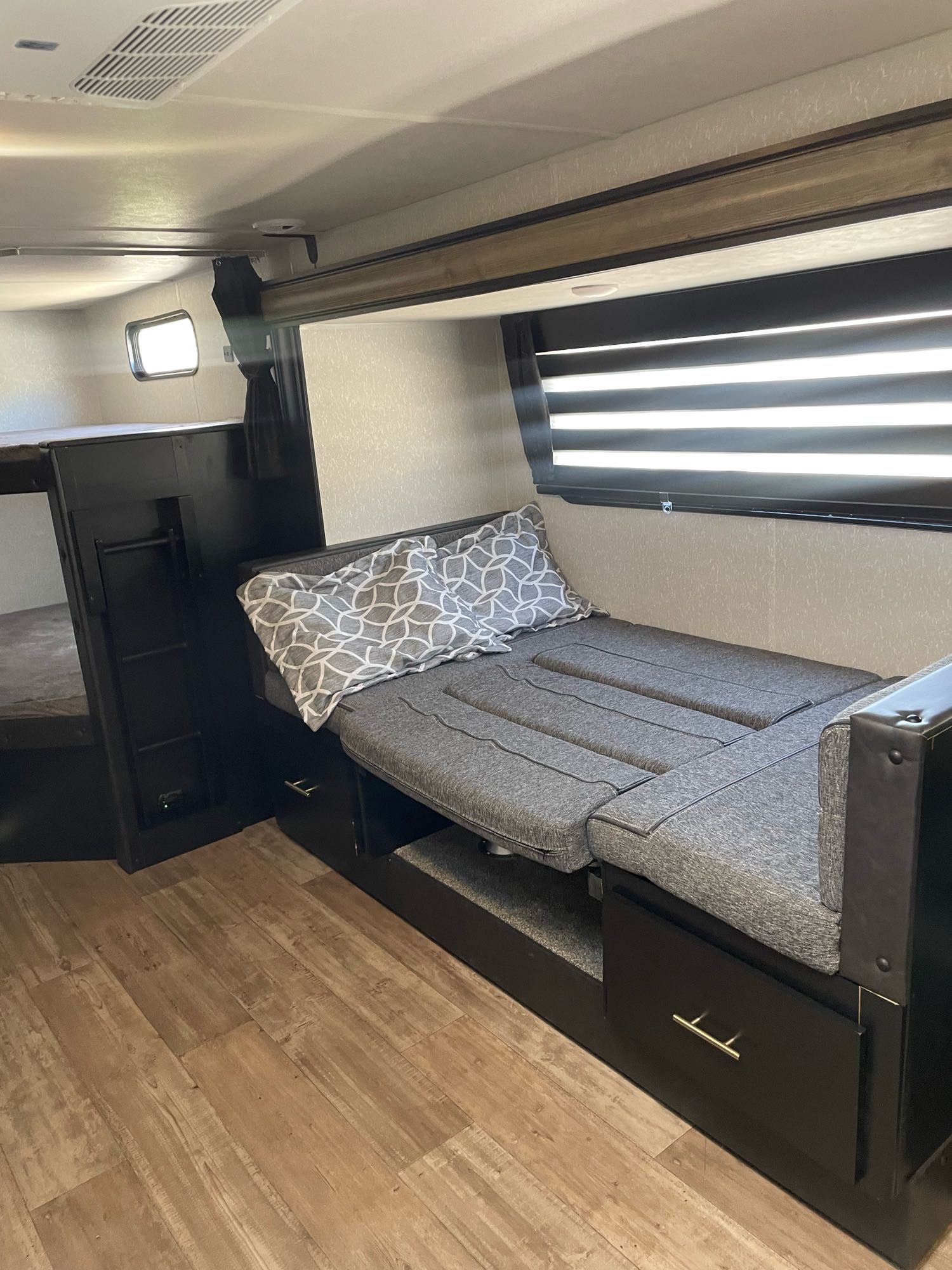 This RV rental interior features a grey convertible sofa with a pillow, bunk beds, wood-look floor, and a window with blinds.. Forest River Cherokee Grey Wolf 2022