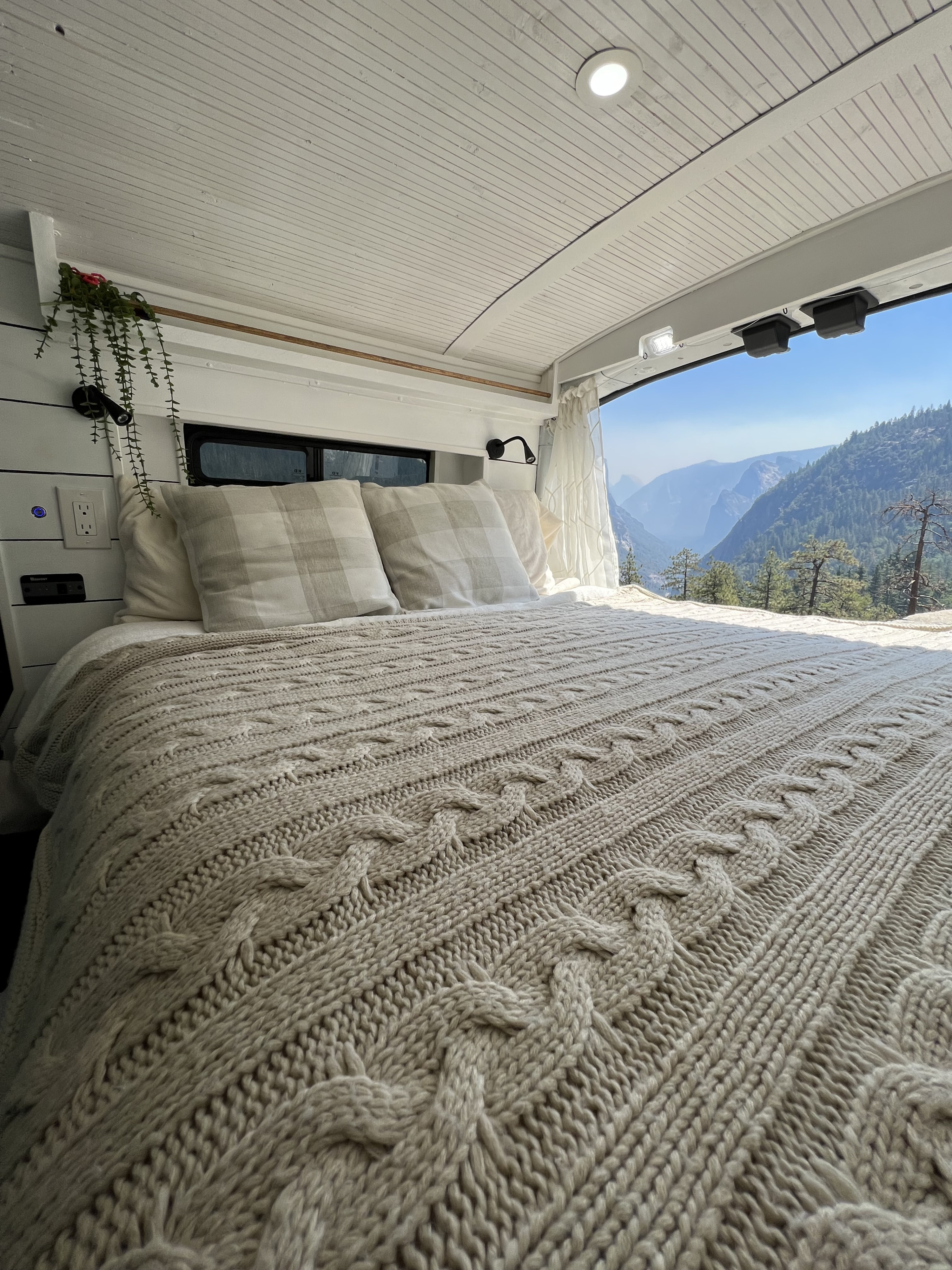 Cozy RV rental bed with a cable knit blanket and mountain view. Perfect for van life adventures, offering comfort in nature.. Ford Transit 2019