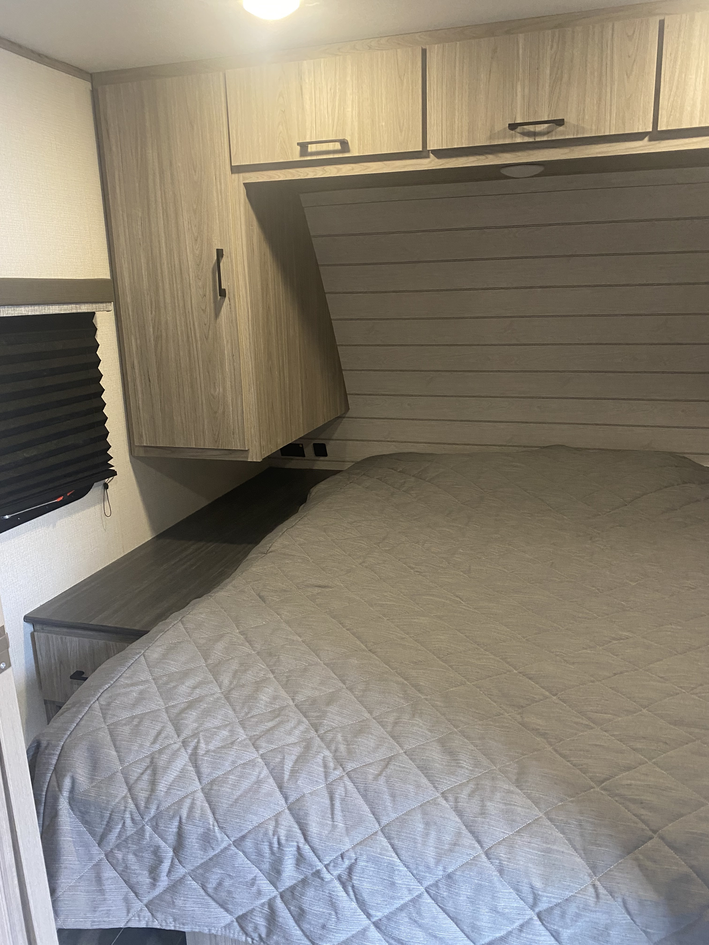 Compact RV rental bedroom interior with a quilted bed, overhead cabinets, and a window blind for a cozy travel experience.. Keystone RV Passport SL 2021