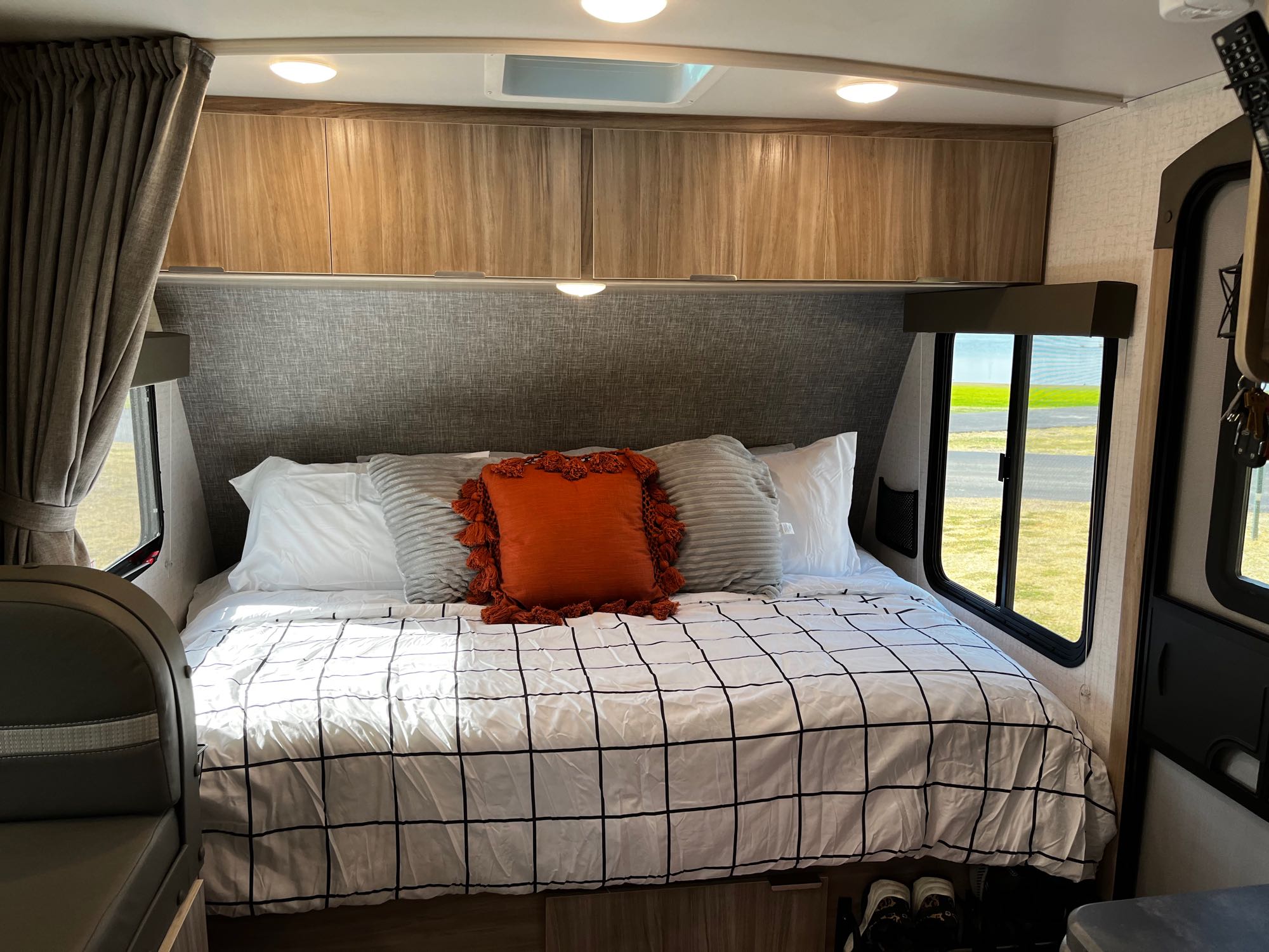 Cozy RV rental bedroom with grid bedding, overhead cabinets, and windows showing green scenery.. Winnebago Micro Minnie 2021